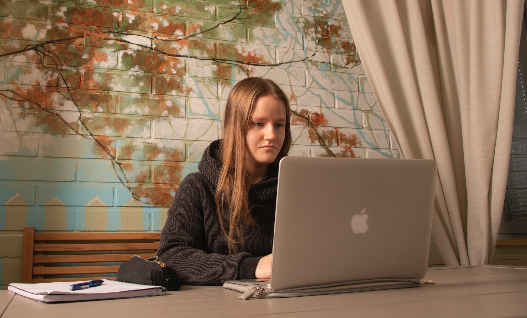 Opiskelija työskentelee tietokoneella ja taustalla näkyy syksyistä puuta esittävä seinämaalaus.