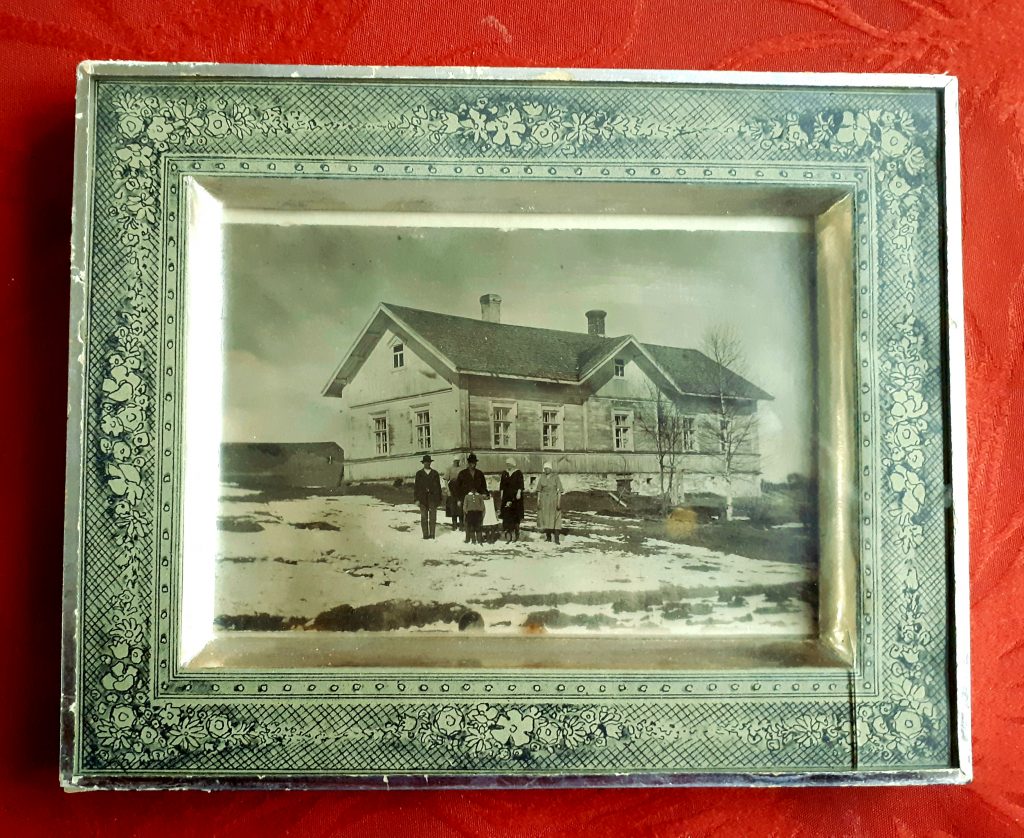 Kehyksissä oleva kuva vanhasta talosta, jonka edustalla seisoo ihmisiä