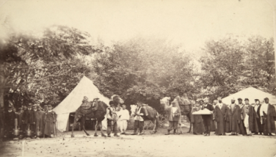 Old photo, behind a tent and in front of it camels and groups of people.