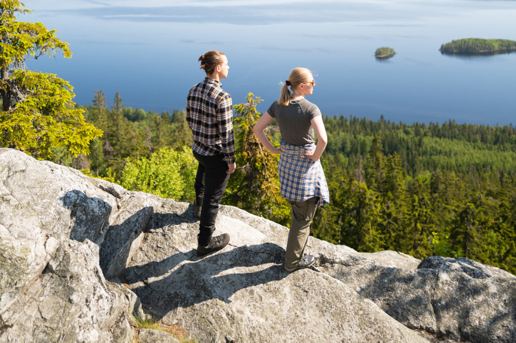Kaksi vaeltajaa seisoo aurinkoisen Kolin laella ja katsoo järvimaisemaa.