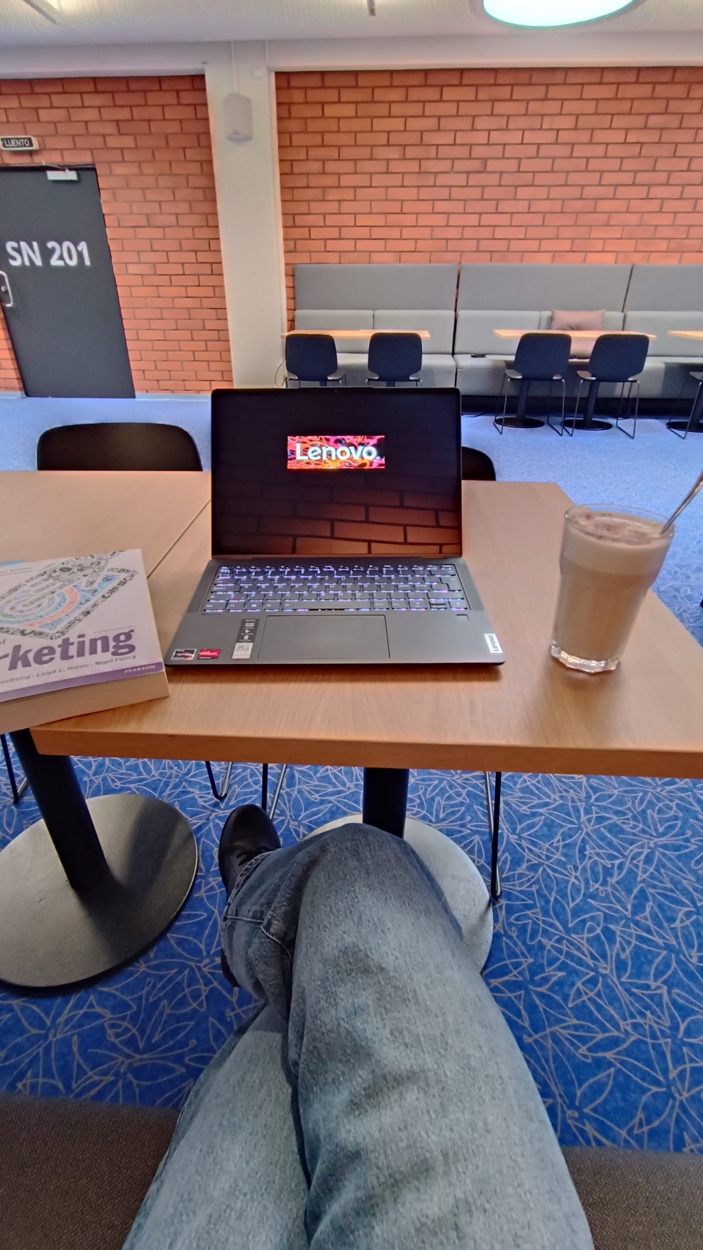 Yliopiston kahvilan pöytä, jossa tietokone, opiskelukirja ja juoma.