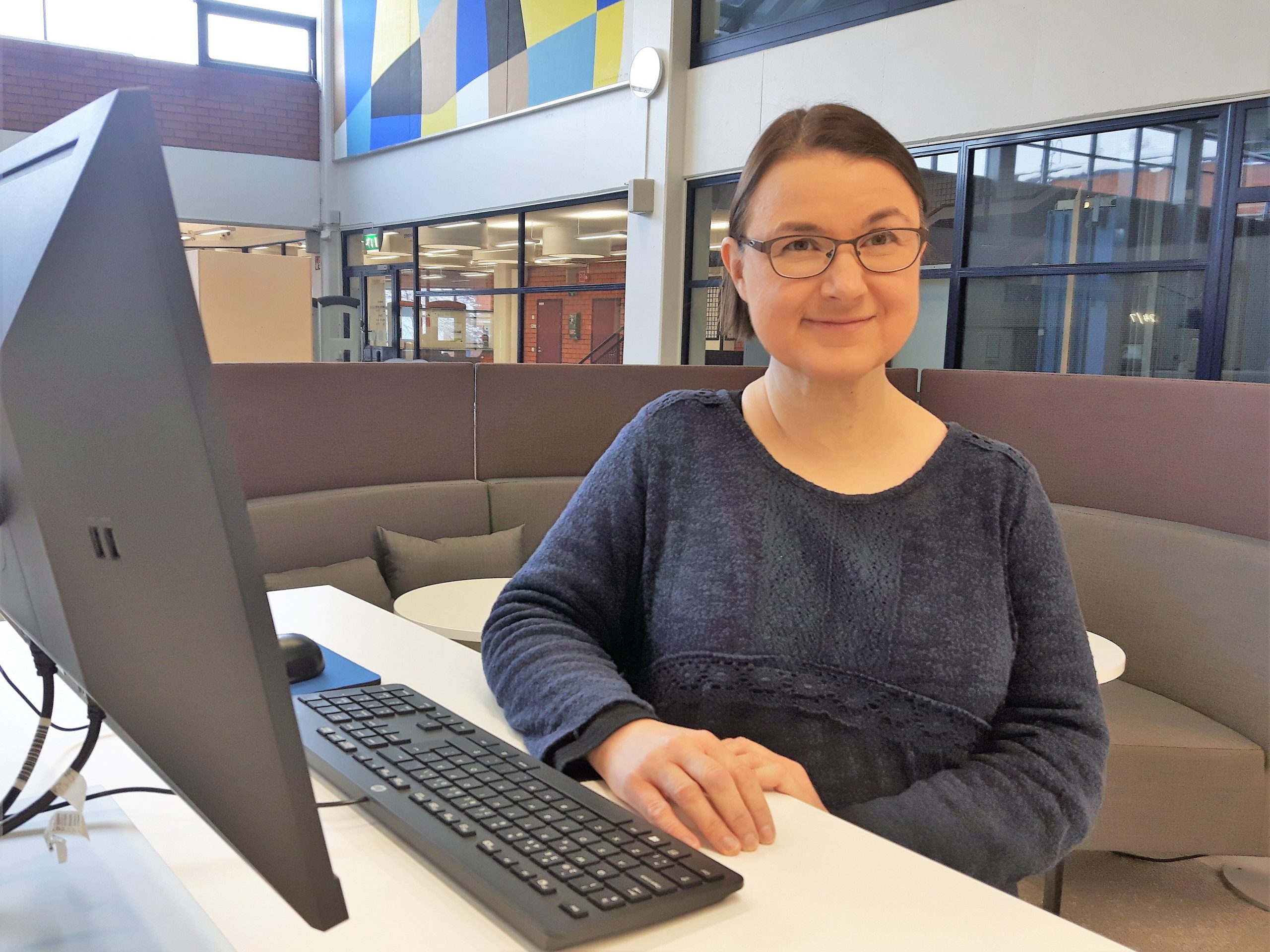 Kirjastossa Katri Rintamäki istuu tietokoneen ääressä, taustalla sohvaryhmä. Katri Rintamäki sitting by a computer, sofas at the background, library interior..