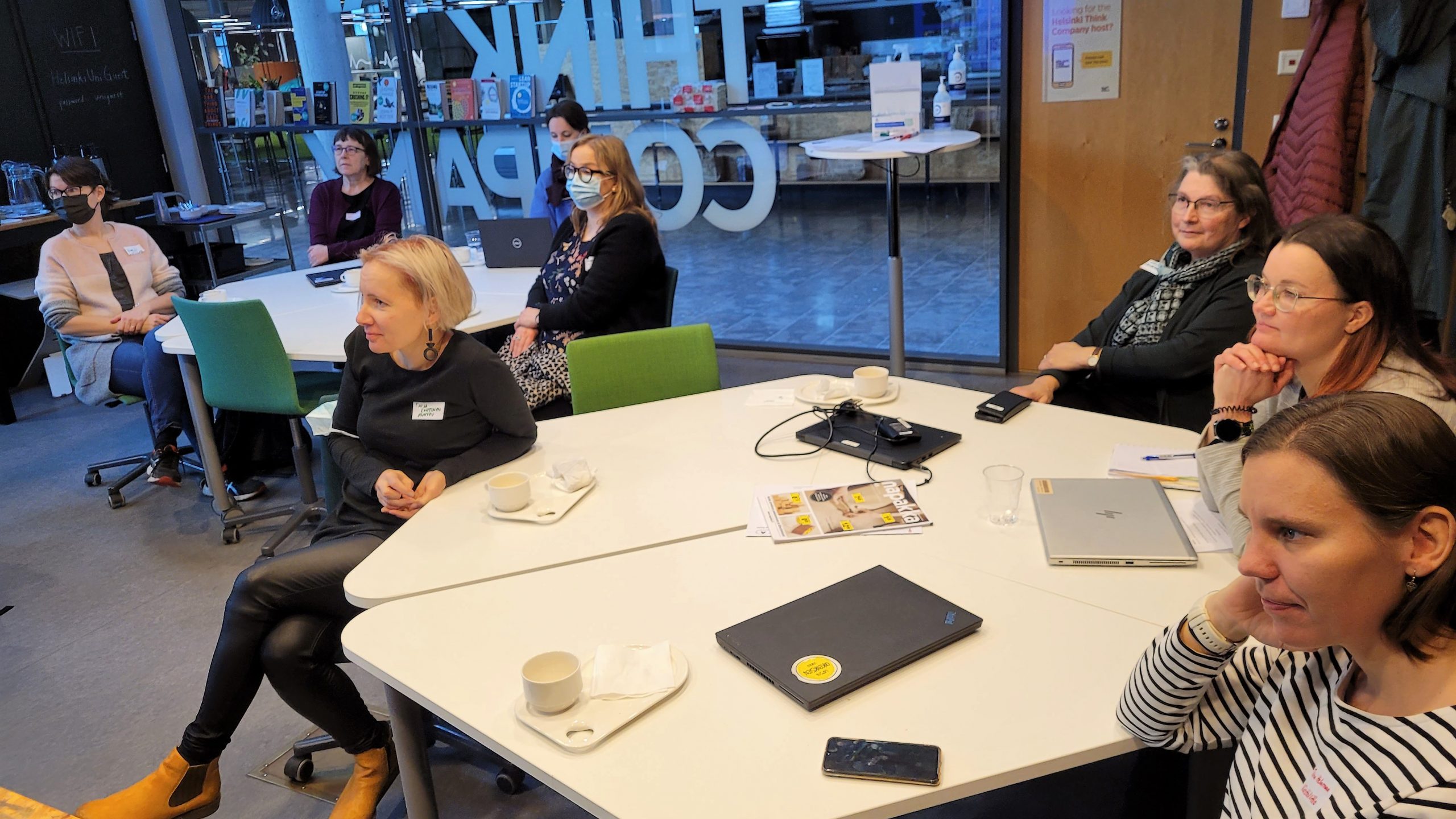 Seminaarissa: ihmisiä, pöytiä joilla kahvikuppeja muistiinpanovälineitä, lasiseinä.| In the seminar: participants, tables, glass wall.