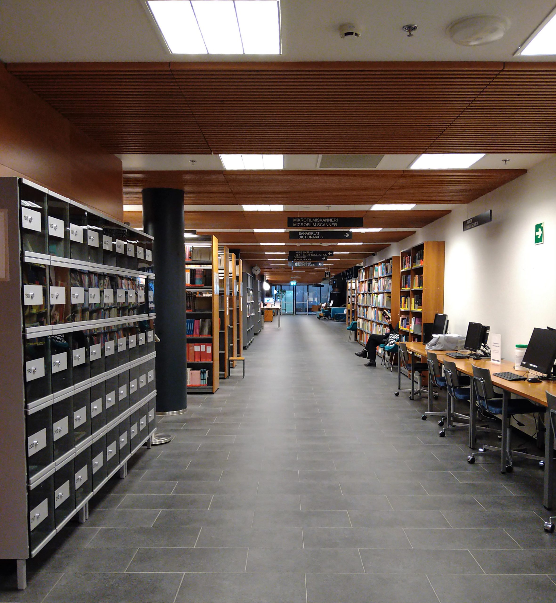 Käytävä, jonka varrella on lokeroita, tietokoneita ja kirjahyllyjä. A passageway with lockers, computers and bookshelves.