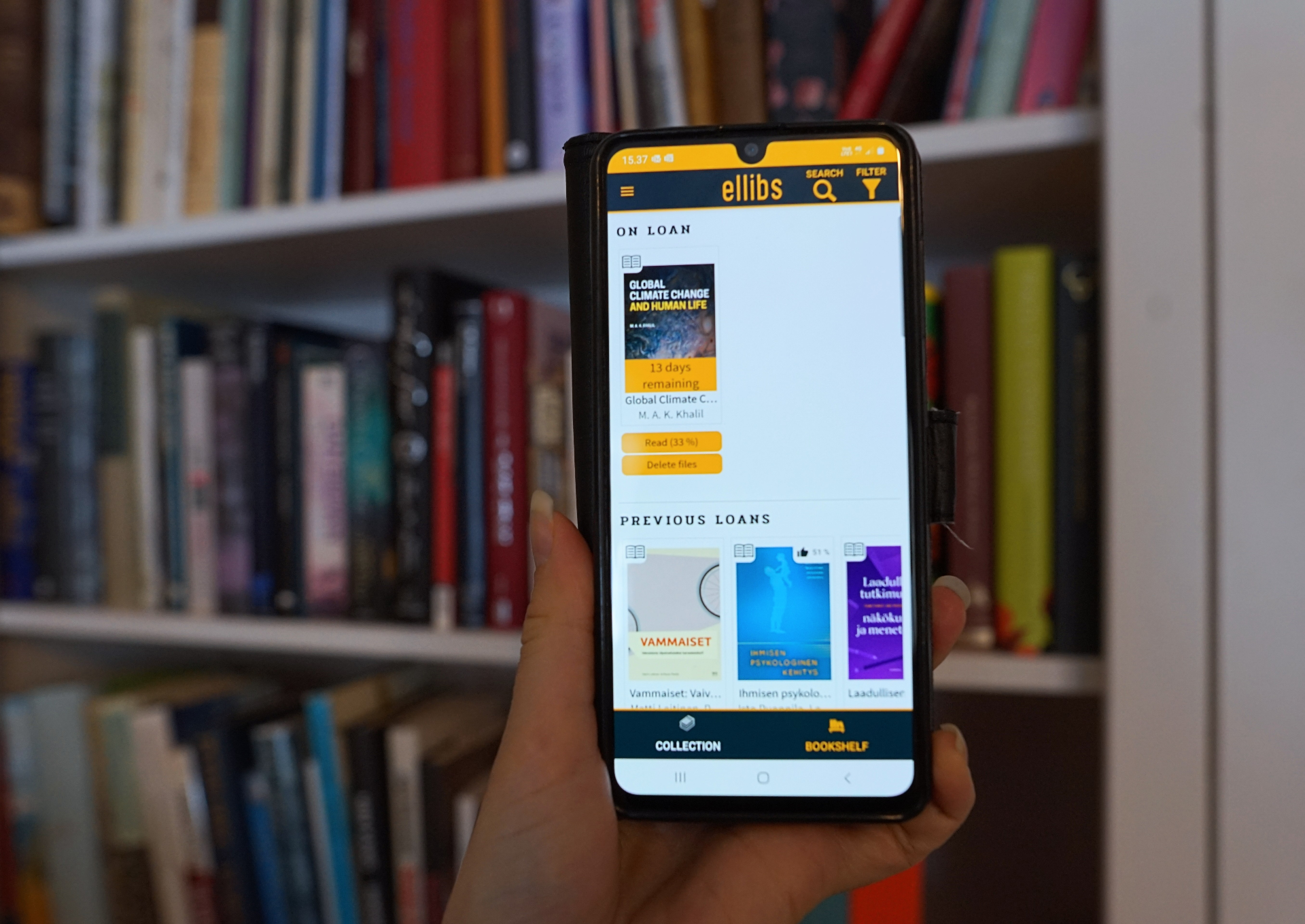 Kuvassa älypuhelin, jossa on auki Ellibs-sovellus. Taustalla kirjahylly. | Smartphone displaying Ellibs app with a bookshelf behind.