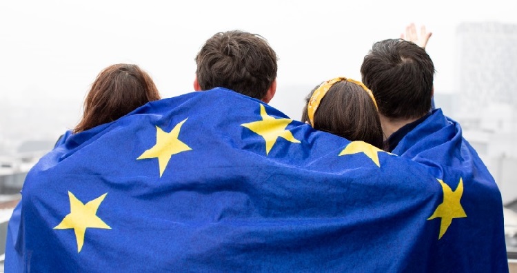 Nuoria ihmisiä takaa päin kuvattuna Euroopan unionin lippu harteillaan. Young people with the European Union flag on their shoulders.