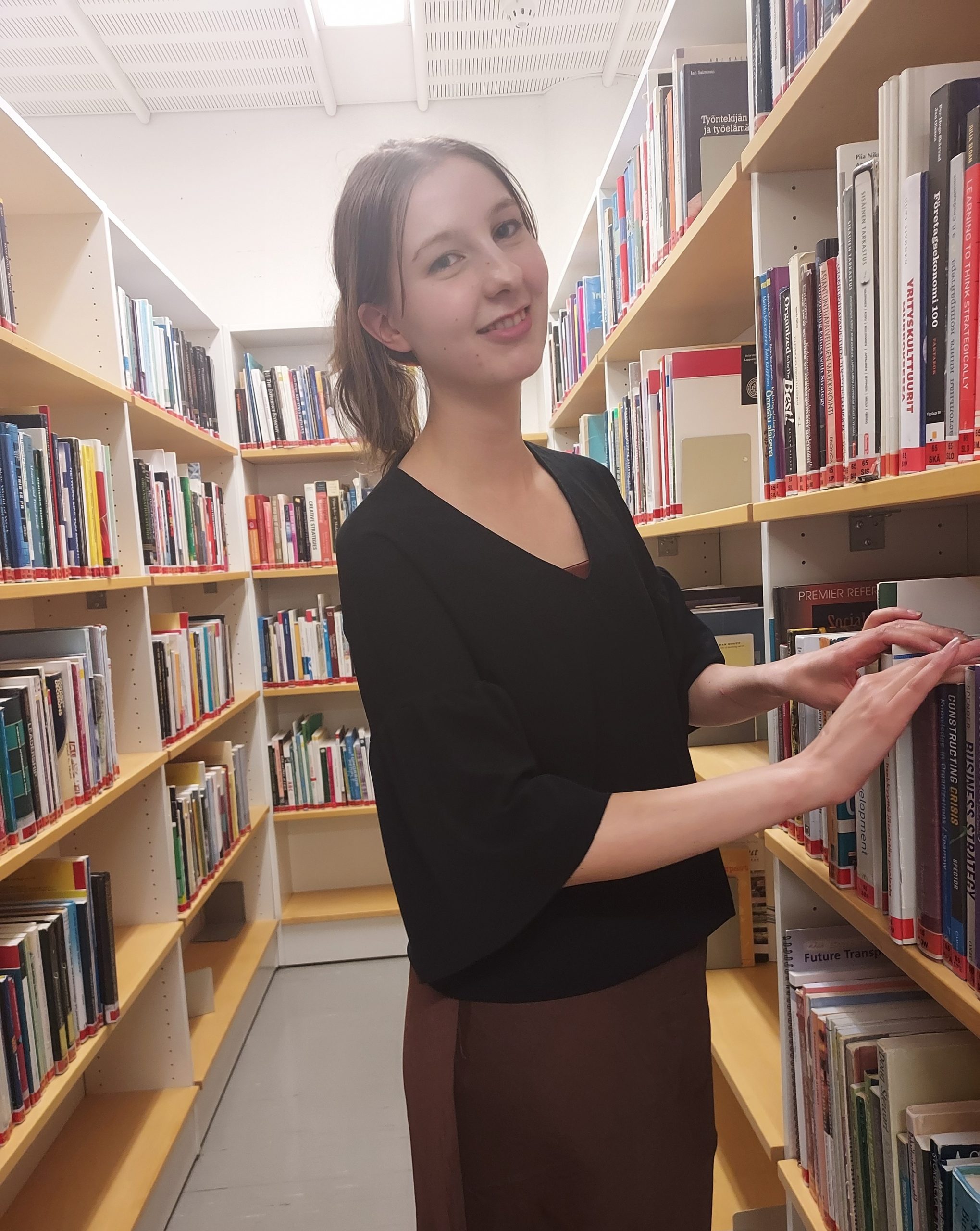 Nainen katsoo kameraan kirjahyllyjen välissä. Woman looking at the camera between book shelves.