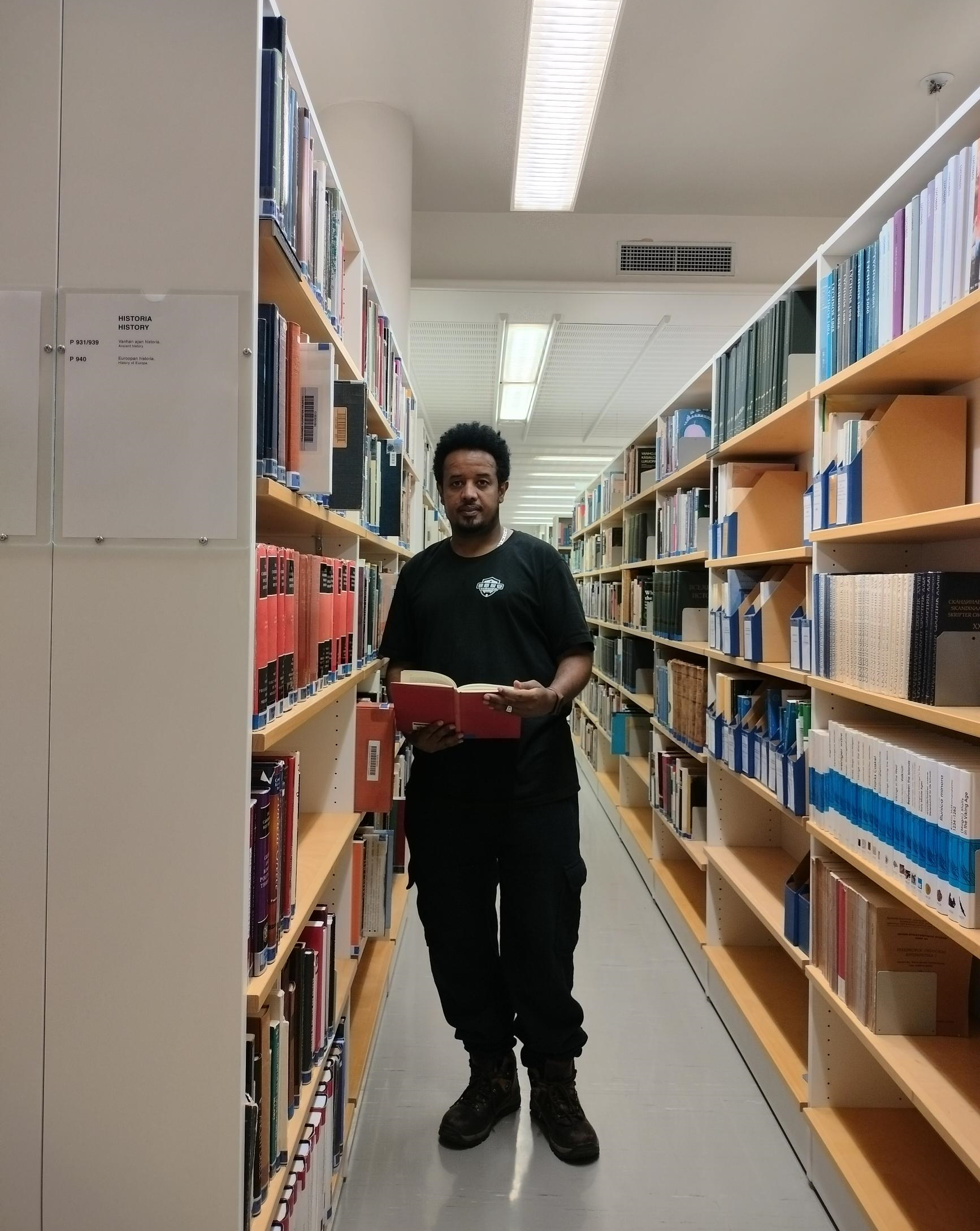 Intern Michael Gebremedhin in a library collection hall. Michael is standing between two shelves and holding an open book in his hands.