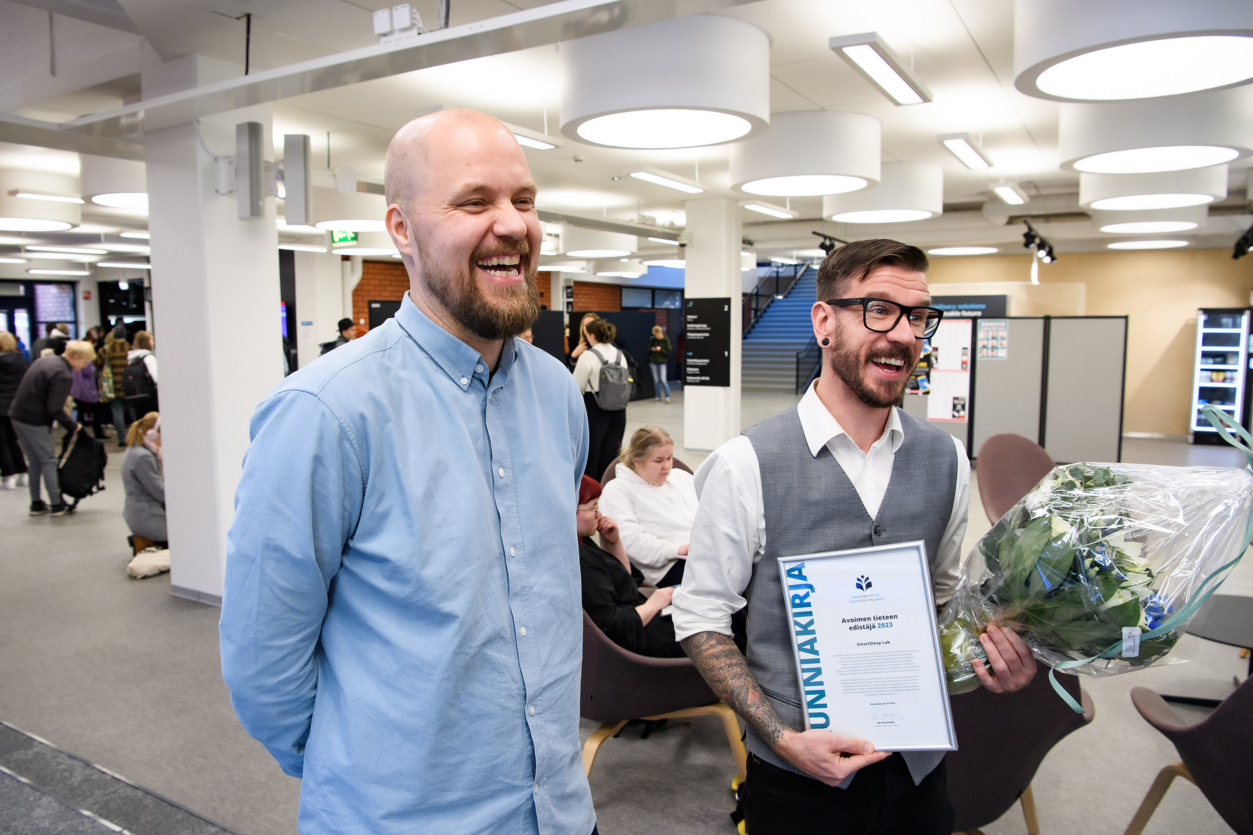 Kaksi nauravaista ihmistä, kukkakimppu, kunniakirja, aulassa. Two people laughing, bucket of flowers, certificate, in a lobby.