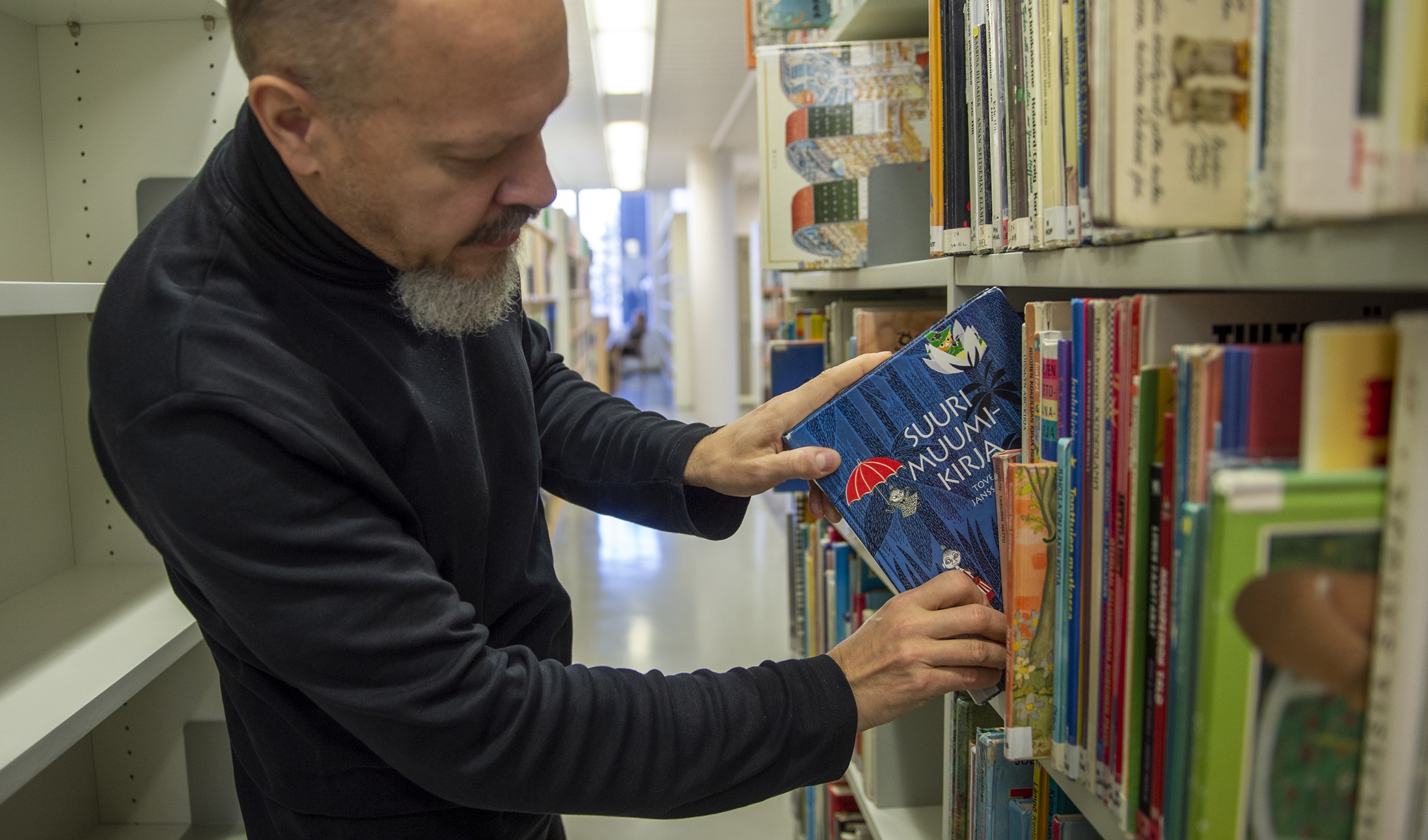 Mies vetämässä esille lastenkirjaa kirjaston hyllystä | A man pulling out a children's book from a library shelf.