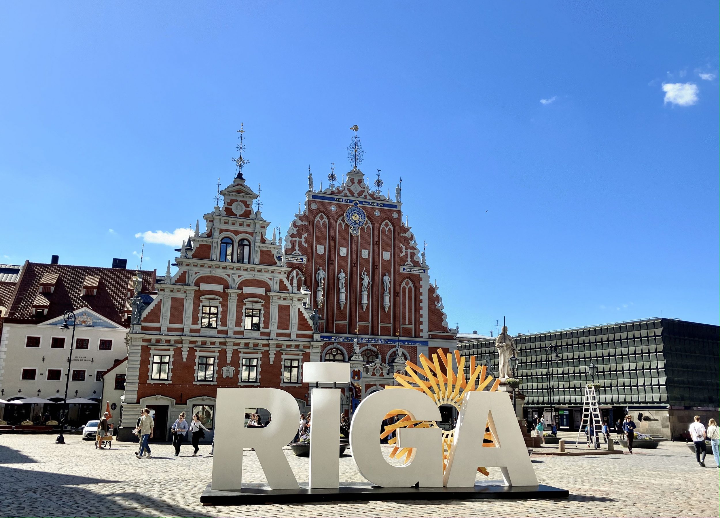 Vanhoja ja uusia rakennuksia aukiolla. Ihmisiä kävelemässä. Patsas, Aurikoinen päivä. Etualalla suurin kirjaimin sana Riga. Old and new buildings on a square. People walking. A statue. Sunny day, In the foreground in capital letters the word Riga.