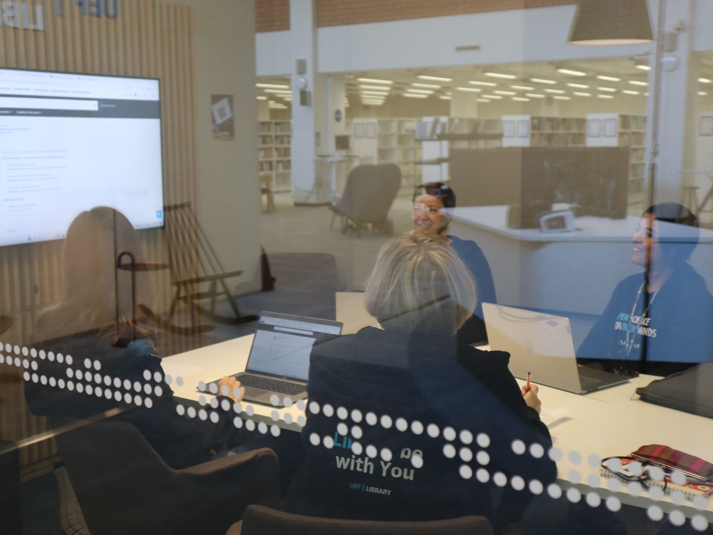 People are sitting around a table, talking and working on laptops. A large screen is visible behind them. The photo is taken from outside the room, through a glass wall.

