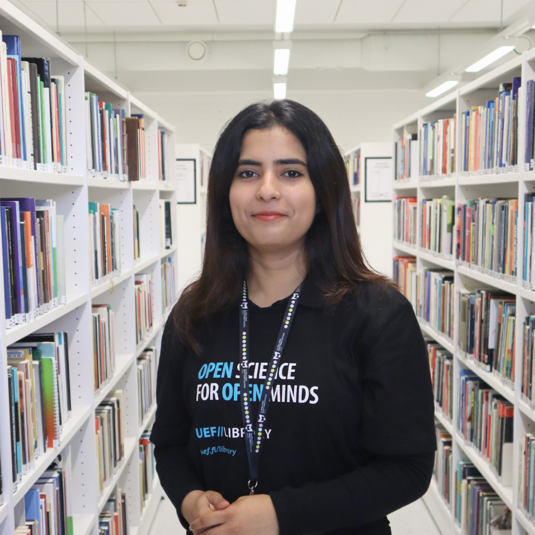 Woman standing in a library aisle surrounded by bookshelves. Nainen seisoo kirjaston käytävällä kirjahyllyjen ympäröimänä.