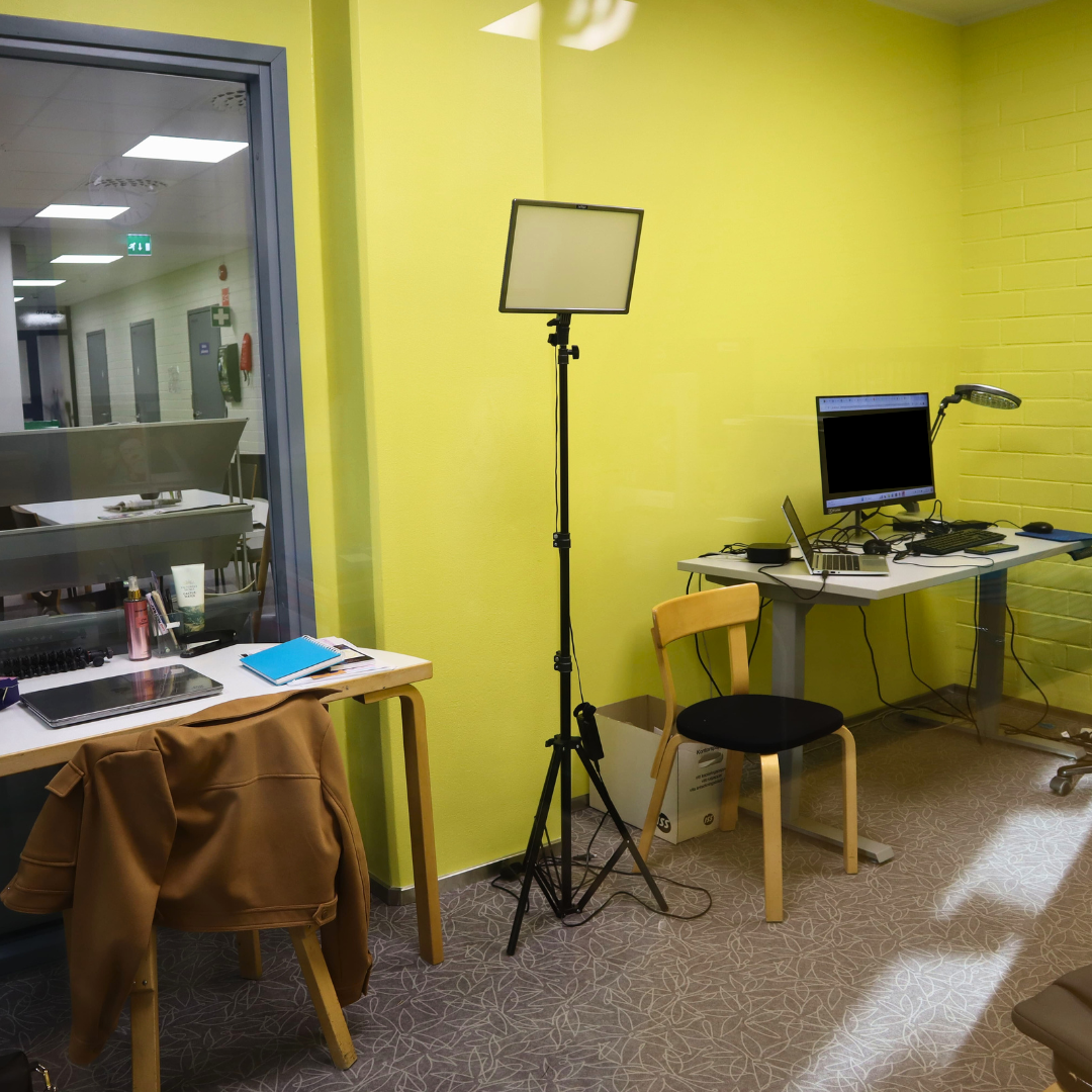 A small, brightly lit room with a yellow wall. A desk in the left with a laptop, notebook, and a brown jacket. A tripod with a light is set up nearby. A computer monitor and a desk lamp are on a table in the right. Pieni, kirkkaasti valaistu huone, jossa on keltainen seinä. Vasemmalla kirjoituspöytä, jossa on kannettava tietokone, vihko ja ruskea takki. Lähistölle on pystytetty kolmijalka, jossa on valo. Tietokoneen monitori ja pöytälamppu ovat oikeanpuoleisella pöydällä.