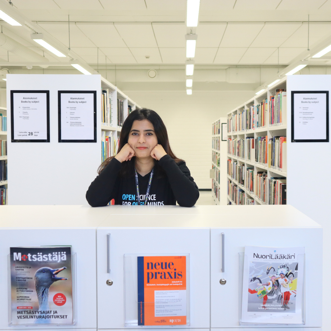 I am standing at a library counter, surrounded by bookshelves, with a gentle smile and hands resting on chin. Wearing a black shirt with 'Open Science for Open Minds' written on it. Magazines displayed in the foreground