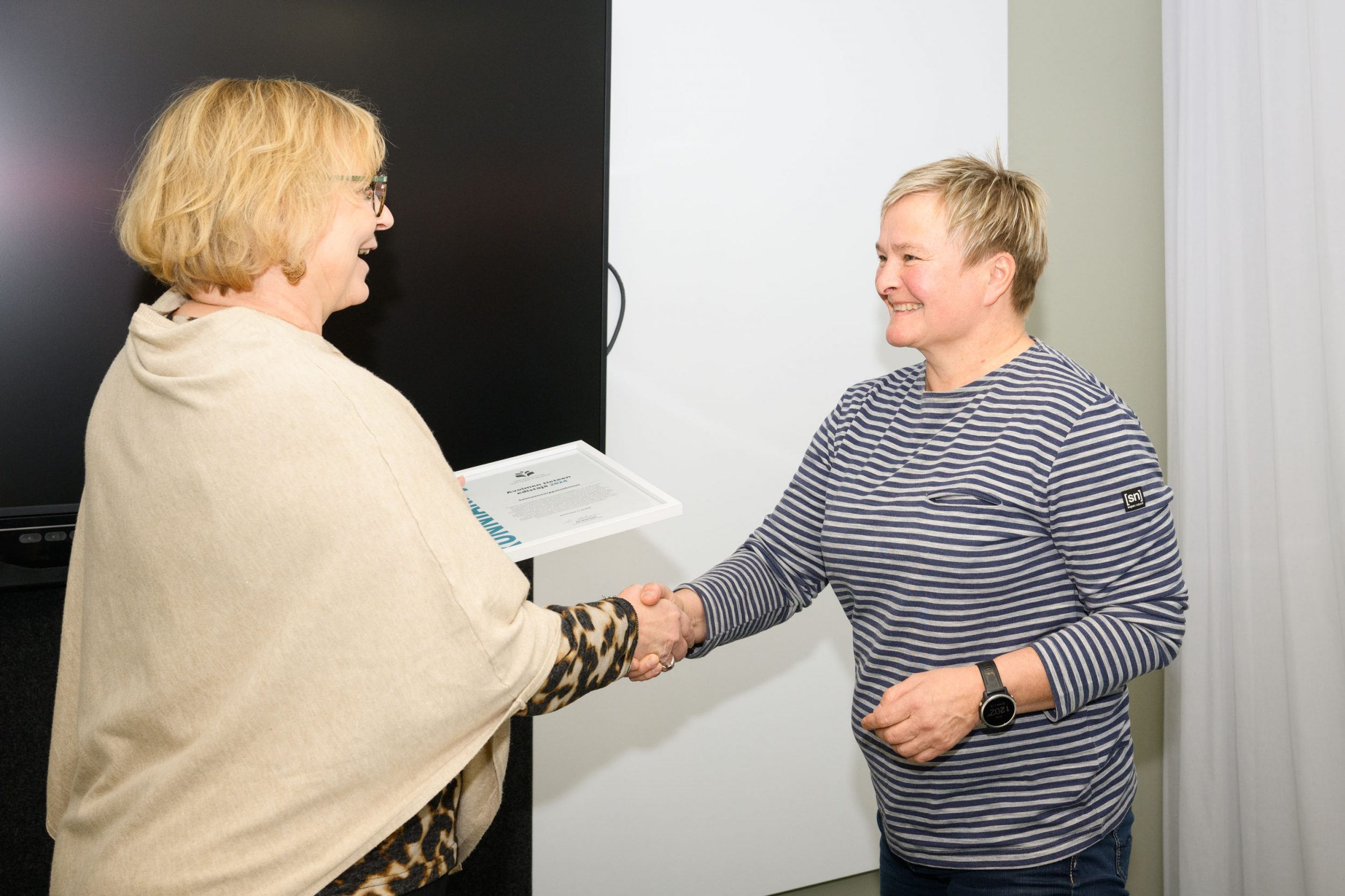 Nainen ojentaa kunniakirjan toiselle naiselle. A woman hands a certificate to another woman.