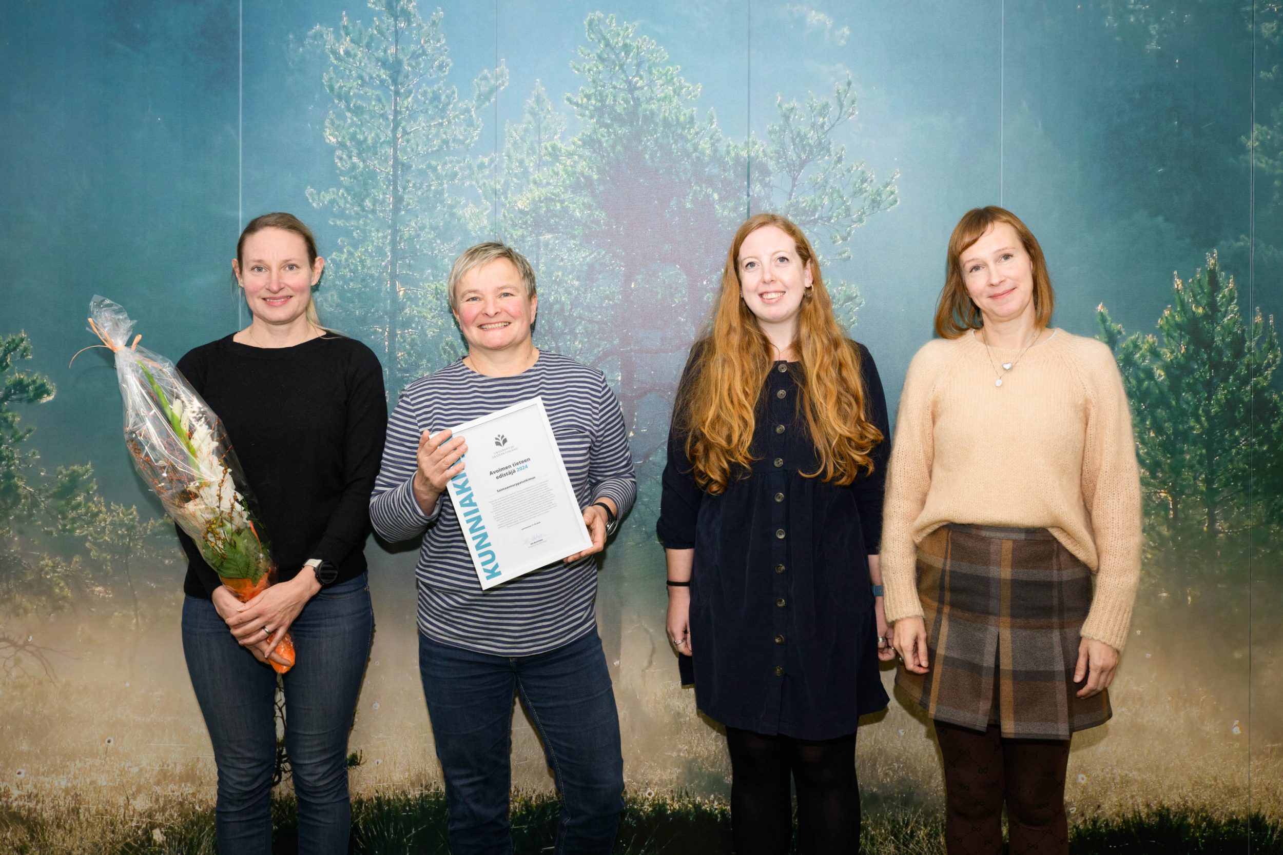 Neljä ihmistä joista yhdellä kukkakimppu ja toisella kunniakirja. Four people, one with a bouquet and the other with a certificate.