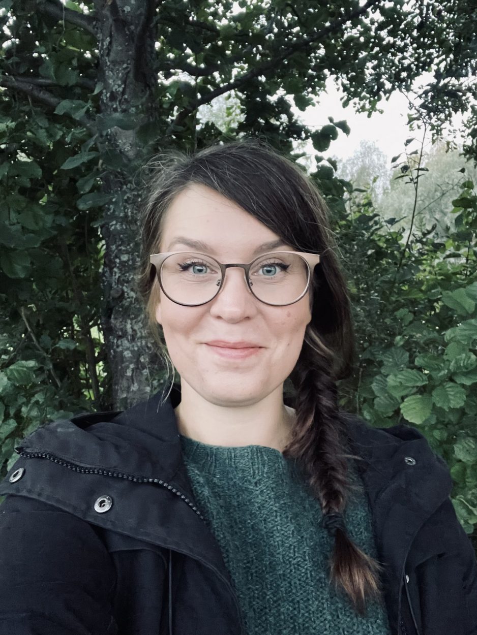 Hymyilevä silmälasipäinen nainen, taustalla puu. A smiling woman with glasses, a tree in the background.