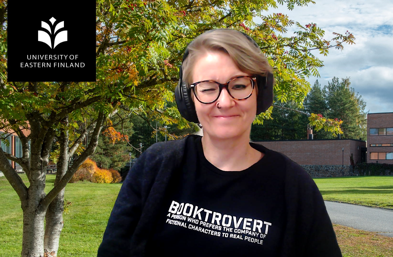 Riitta istuu kuulokkeet korvillaan musiikkia kuunnellen ja iskee silmää. Taustalla näkyy syksyinen Joensuun kampus. | Riitta sitting with her headphones on while listening to music and winking. In the background Joensuu campus in the autumn.