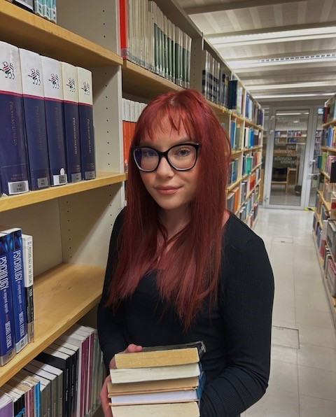 kirjasto, kirjahyllyjä, nainen jolla on kirjapino käsissään. Library, bookshelves, woman holding a pile of books.