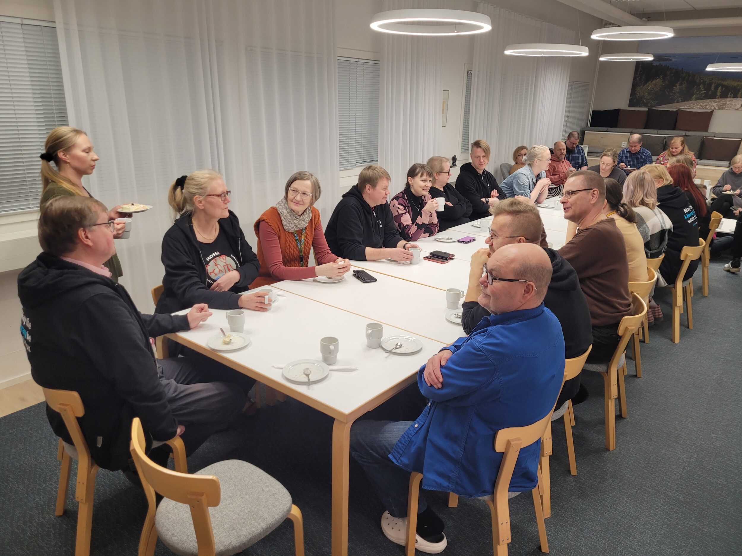 Suuri joukko ihmisiä kahvipöydän ääressä. | Large group of people around a coffee table.