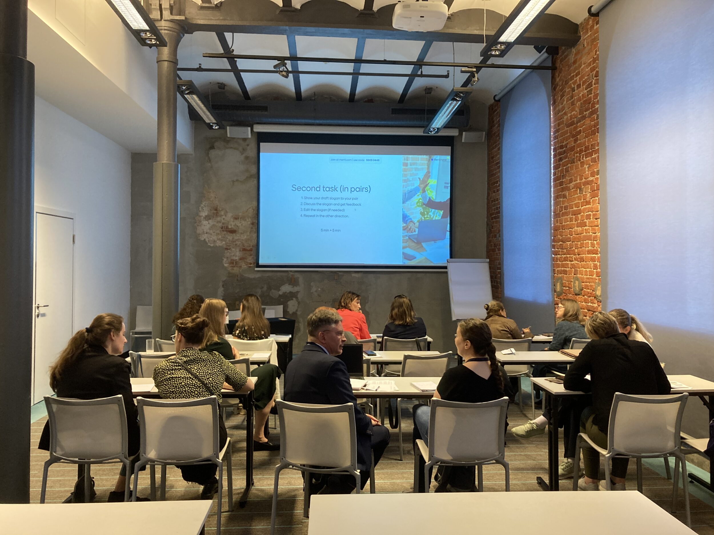 Tiiliseinäinen seminaarisali, ihmisiä juttelemassa pareittain, taustalla valkokankaalla parityön ohjeistus. A brick-walled seminar hall, people talking in pairs, in the background instructions for pair work.