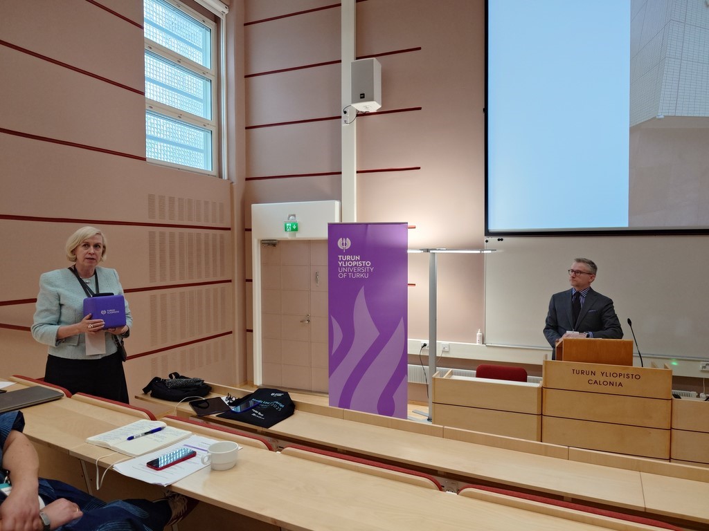 Turun yliopiston kirjastonjohtaja Heli Kautonen seisomassa auditorion penkkien välissä ja professori Janne Salminen seisomassa puhujapöydän takana. Library Director Heli Kautonen of Turku University Library and professor Janne Salminen.