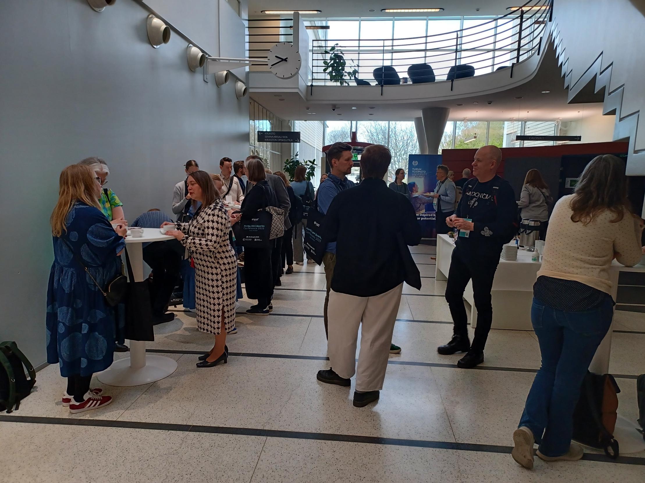 Konferenssin osallistujat keskustelevat keskenään ja juovat kahvia tai teetä. Kaikki seisovat, osa korkeiden pöytien ääressä. Participants of the conference discussing and drinking coffee or tea.