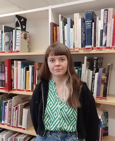 Milja seisoo kirjahyllyn edessä kirjastossa. Milja stands in front of a bookshelf in library.