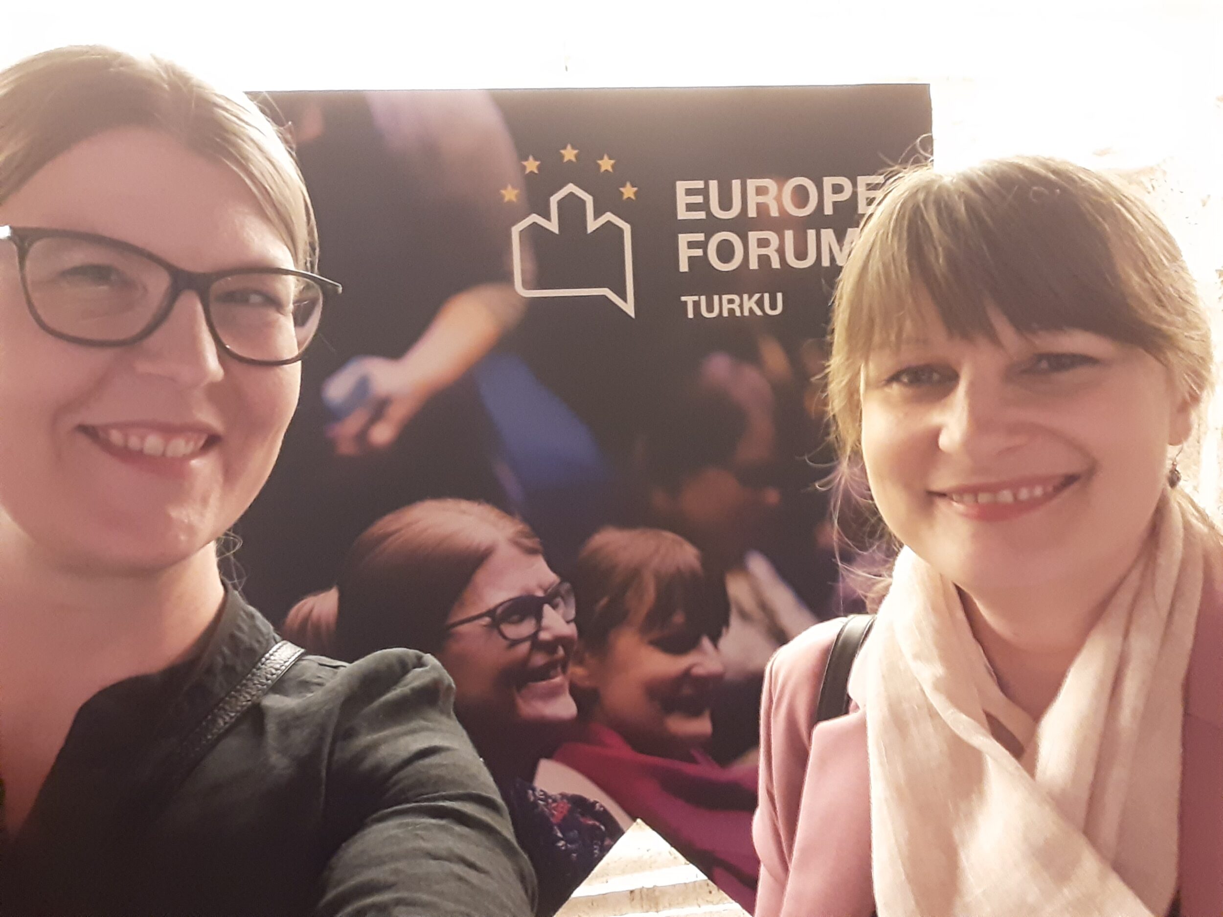 Taustalla Eurooppa-foorumin mainosjuliste, jossa yleisön joukosta erottuvat hyvin kahden henkilön kasvot. Samat henkilöt poseeraavat julisteen edessä. In the background is a poster for the Europe Forum, where the faces of two people stand out well from the audience. The same people pose in front of the poster.