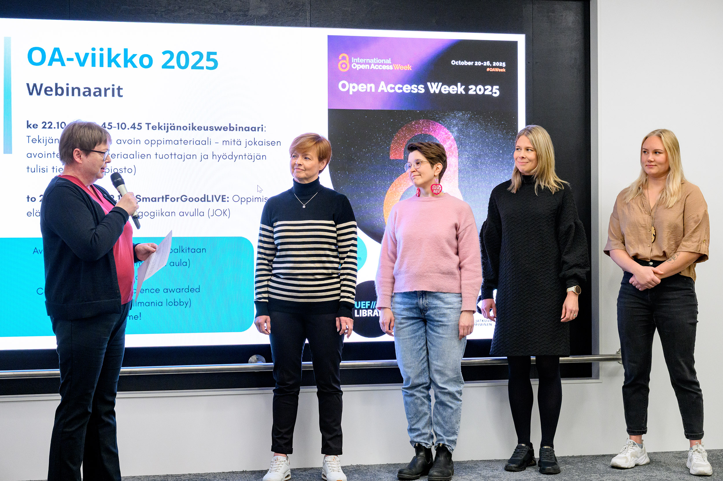 Lavalla viisi henkilöä, joista yksi puhuu mikrofoniin. | On stage, five people, one speaking to a microphone.