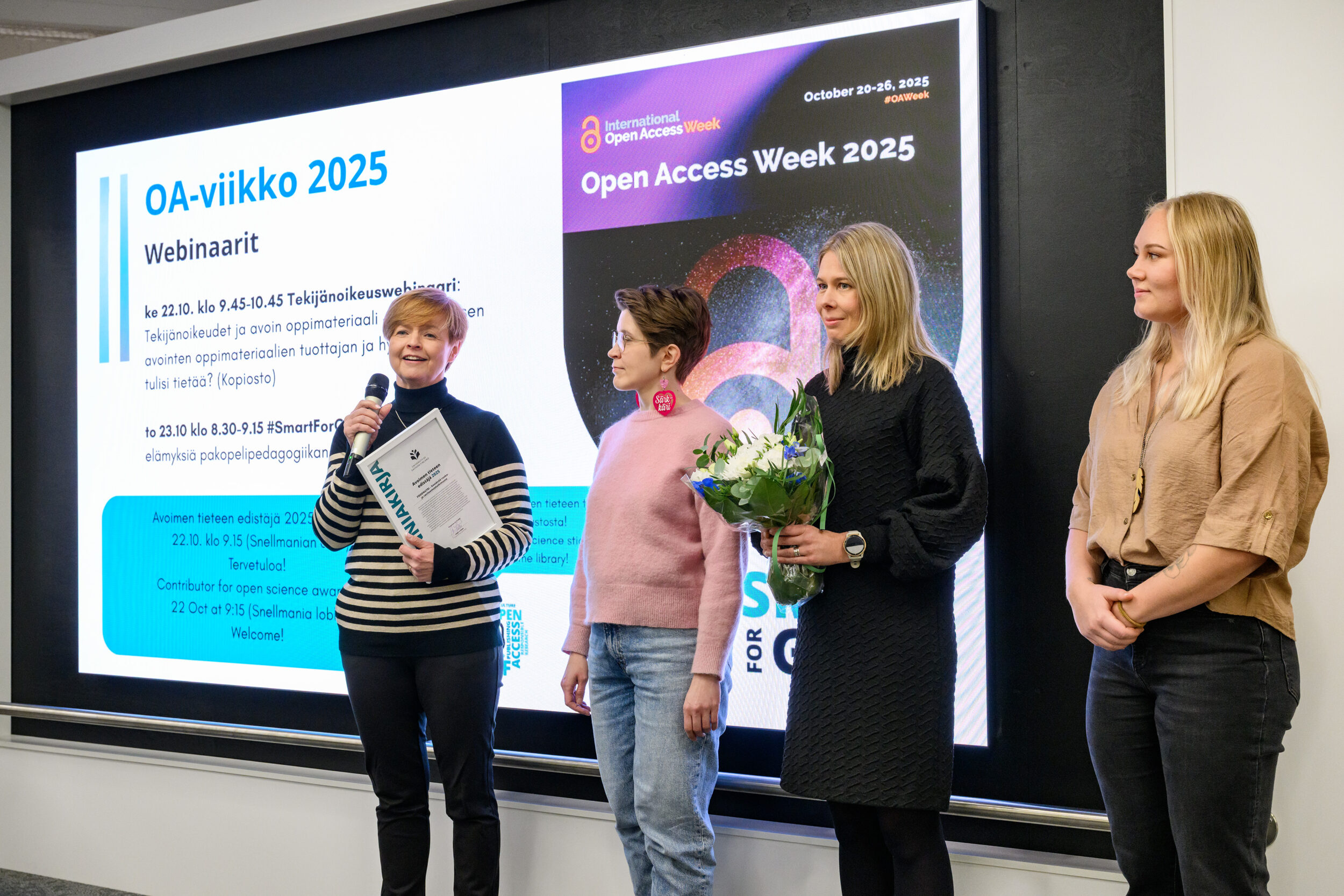 Neljä ihmistä lavalla. Yhdellä mikrofoni ja kunniakirja, toisella kukkakimppu. On stage four people. One holding a microphone and a certificate, another one a bucket of flowers.
