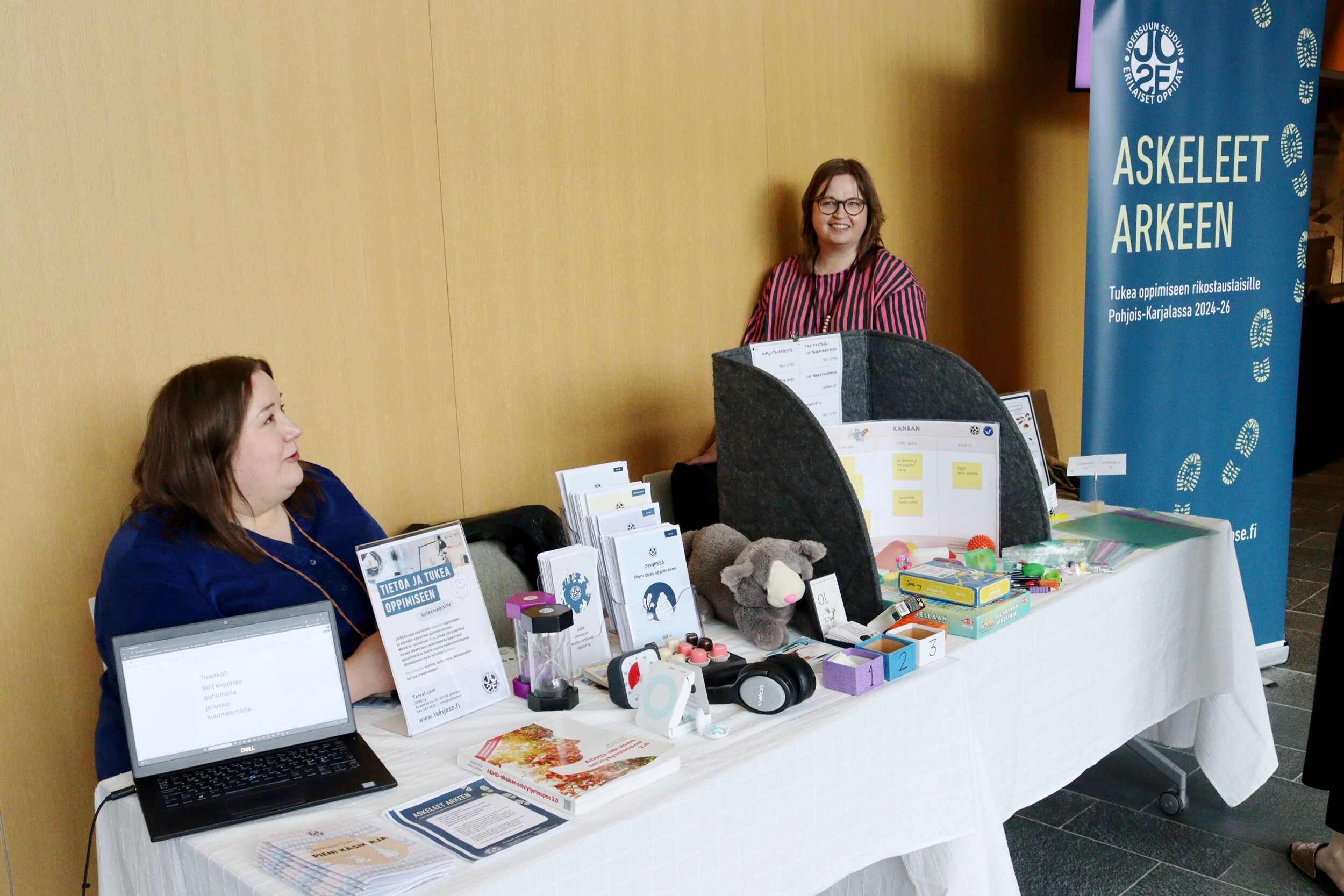 Kaksi henkilöä esittelee Joensuun seudun erilaiset oppijat ry:n infopistettä. Two persons tell about the info booth of Joensuu Regional Association for Diverse Learners.