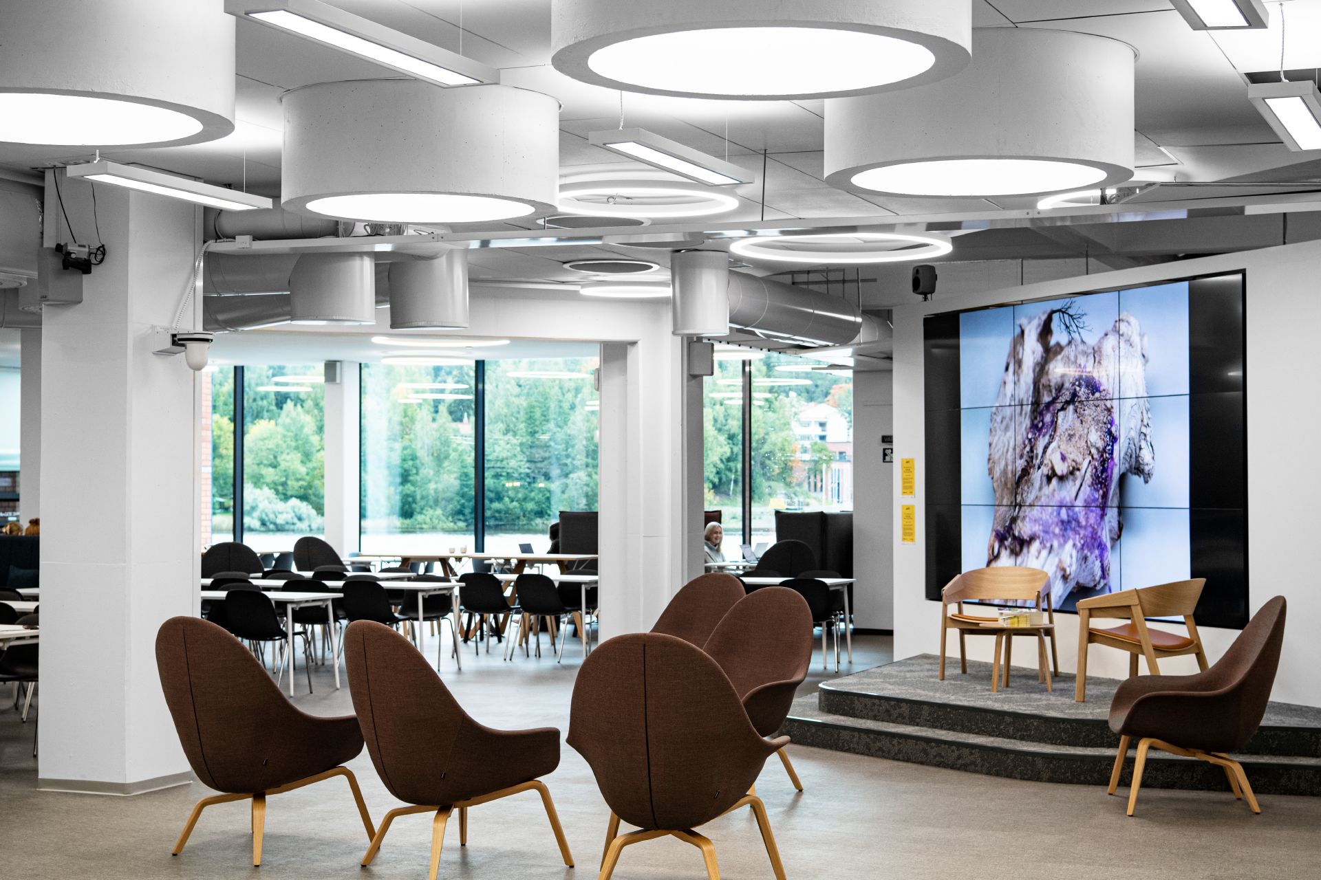 Snellmania-rakennuksen aulassa on tuoleja, pöytiä, pieni esiintymislava ja iso infonäyttö. | There are chairs, tables, a small stage and a large info screen in the lobby of the Snellmania building.