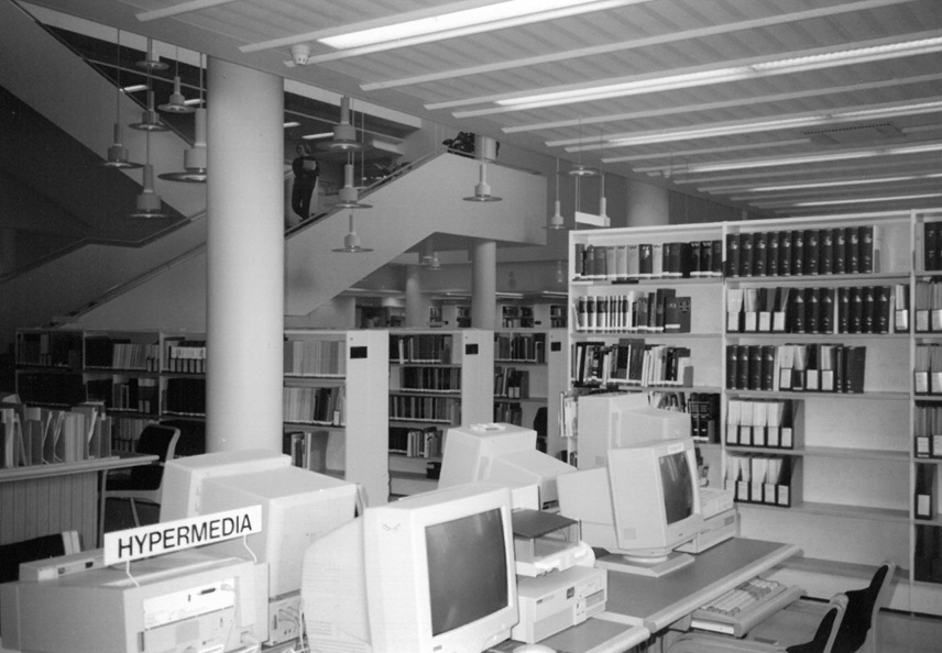 Computers on desks, with reference works on bookshelves in the background.