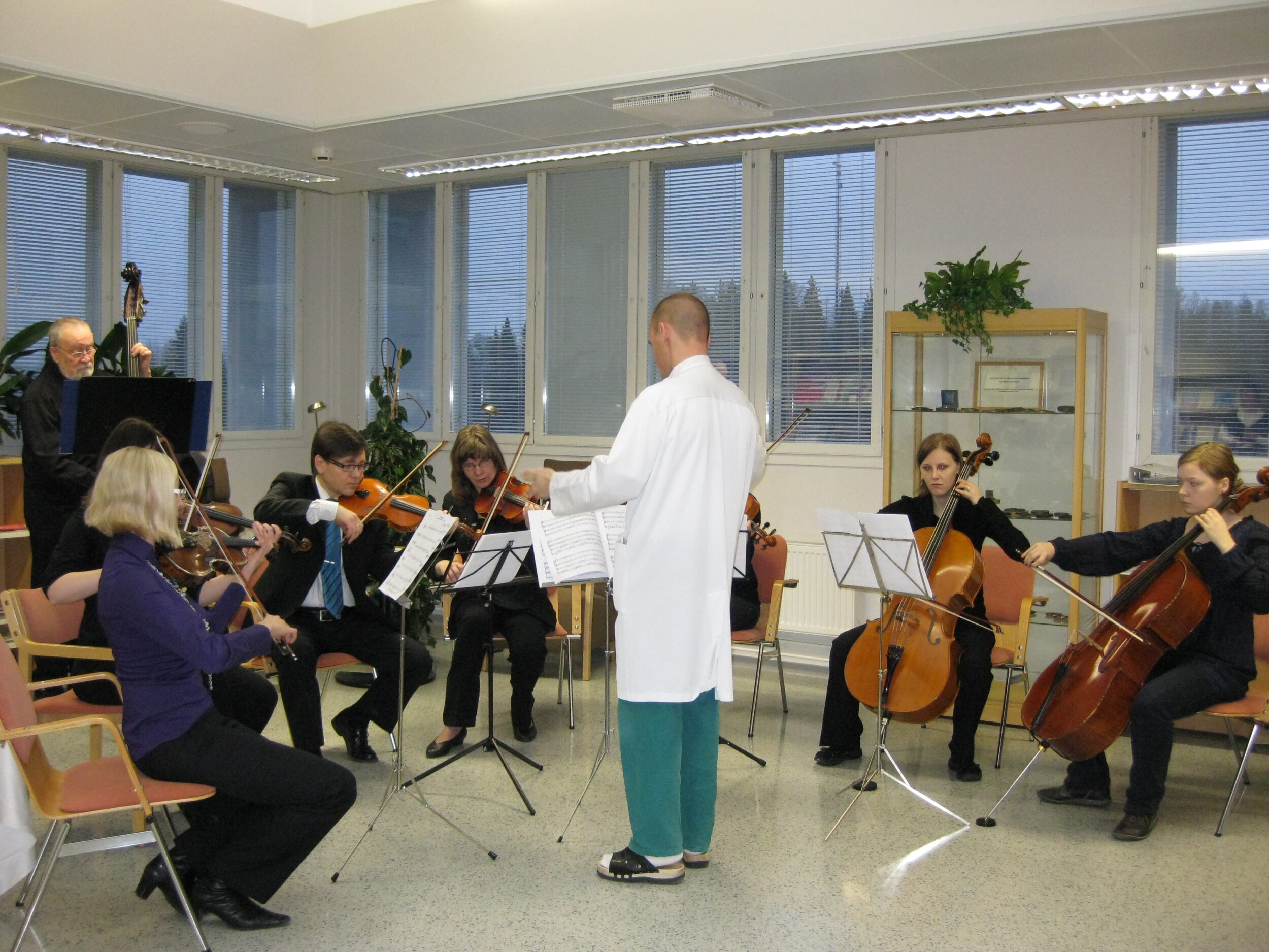 Jousisoitiorkesteri soittaa. Orkesterinjohtajalla on yllään valkoinen lääkärintakki. 