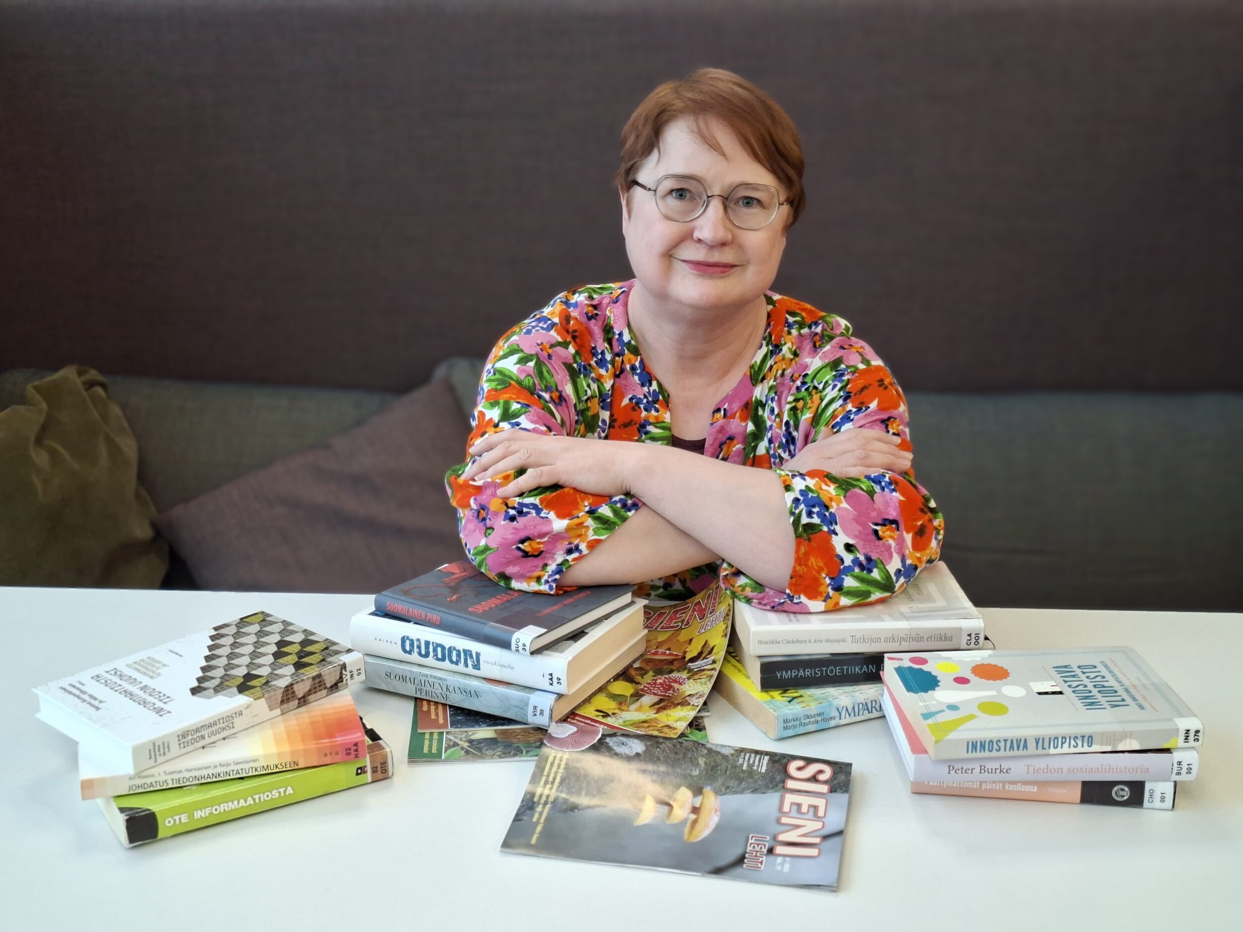 Hymyilevä nainen, jolla on monivärinen paita, istuu harmaalla sohvalla. Hänen edessään on kirjoja ja lehtiä pöydällä, ja hän on asettanut kyynärpäänsä kahdelle matalalle kirjapinolle. Henkilön vieressä on kaksi sohvatyynyä. Taustalla on tumma kangas. A smiling woman wearing a multicolored shirt is sitting on a gray sofa. In front of her, on the table, there are books and magazines, and she has placed her elbows on two low stacks of books. Next to her are two sofa cushions. In the background, there is a dark fabric.
