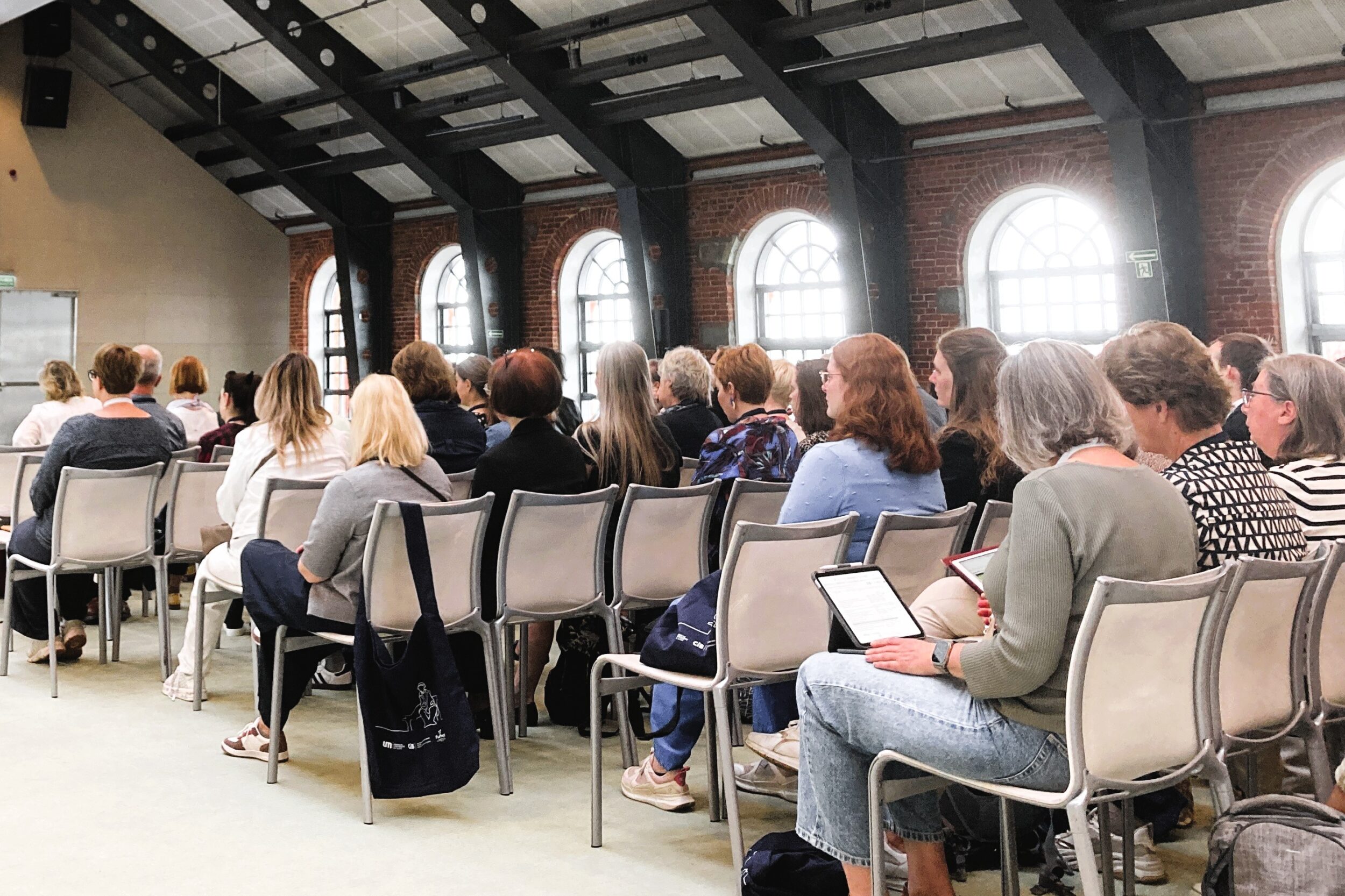 Valoisa luentosali, jossa tiiliseinä, suuret ikkunat ja katto korkealla. Ihmisiä kuvattuna takaapäin istumassa valkoisilla tuoleilla kuuntelemassa esitystä ja tekemässä muistiinpanoja. Bright lecture hall with brick wall, large windows and high roof. People shown from behind sitting in white chairs listening to the presentations and taking notes.
