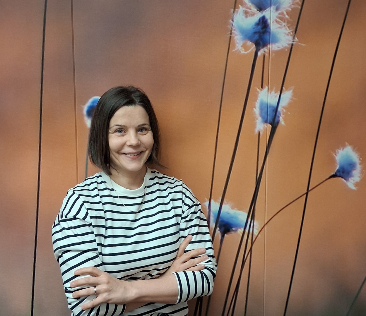 Raitapaitainen nainen hymyilee kädet ristissä seinän edessä.
A woman in a striped shirt smiles with her arms crossed in front of a wall.
