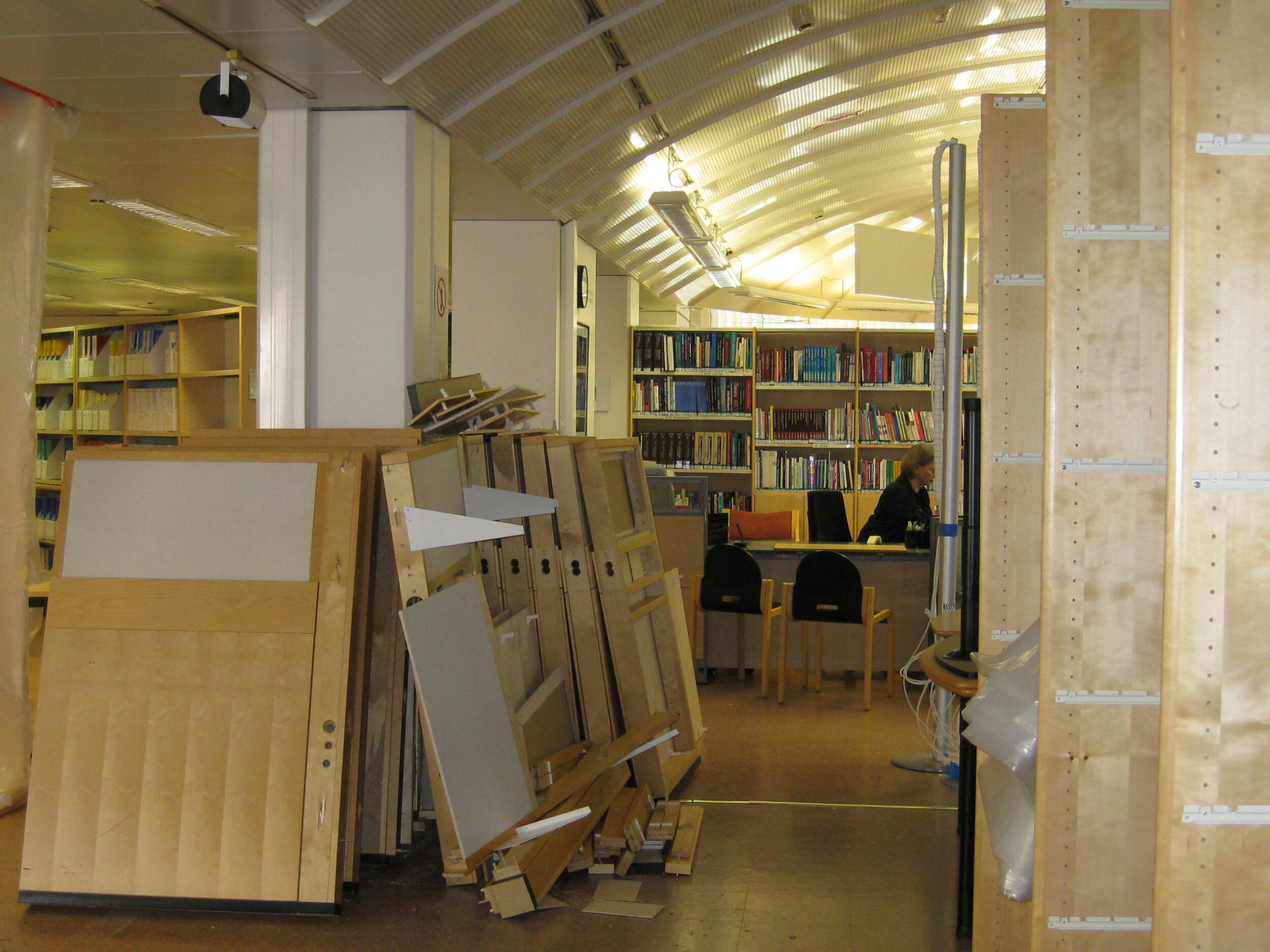 Taustalla kirjahylly kirjoineen ja kirjaston palvelupiste, etualalla tyhjiä hyllyjä ja kasassa muita huonekaluja. 