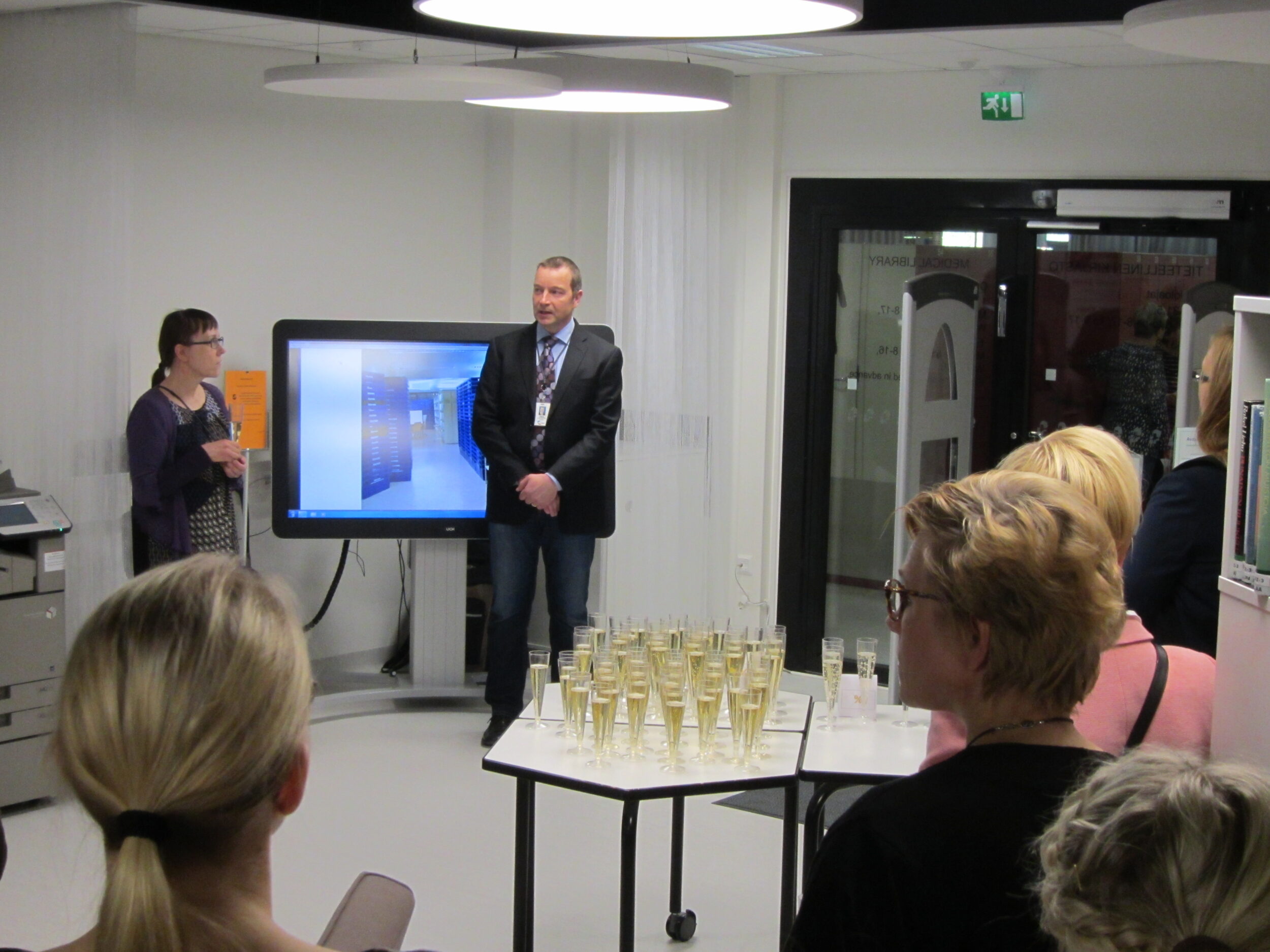 Ihmisiä seisomassa, pöydällä kuohuviinilaseja. 