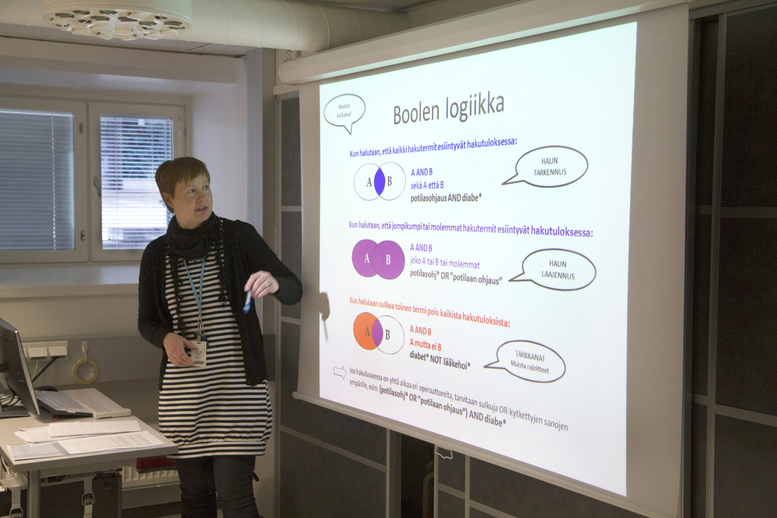 Opetustilanne, jossa henkilö seisoon tietokonepöydän ja valkokankaan välissä. Valkokankaalla esitys Boolen logiikasta.