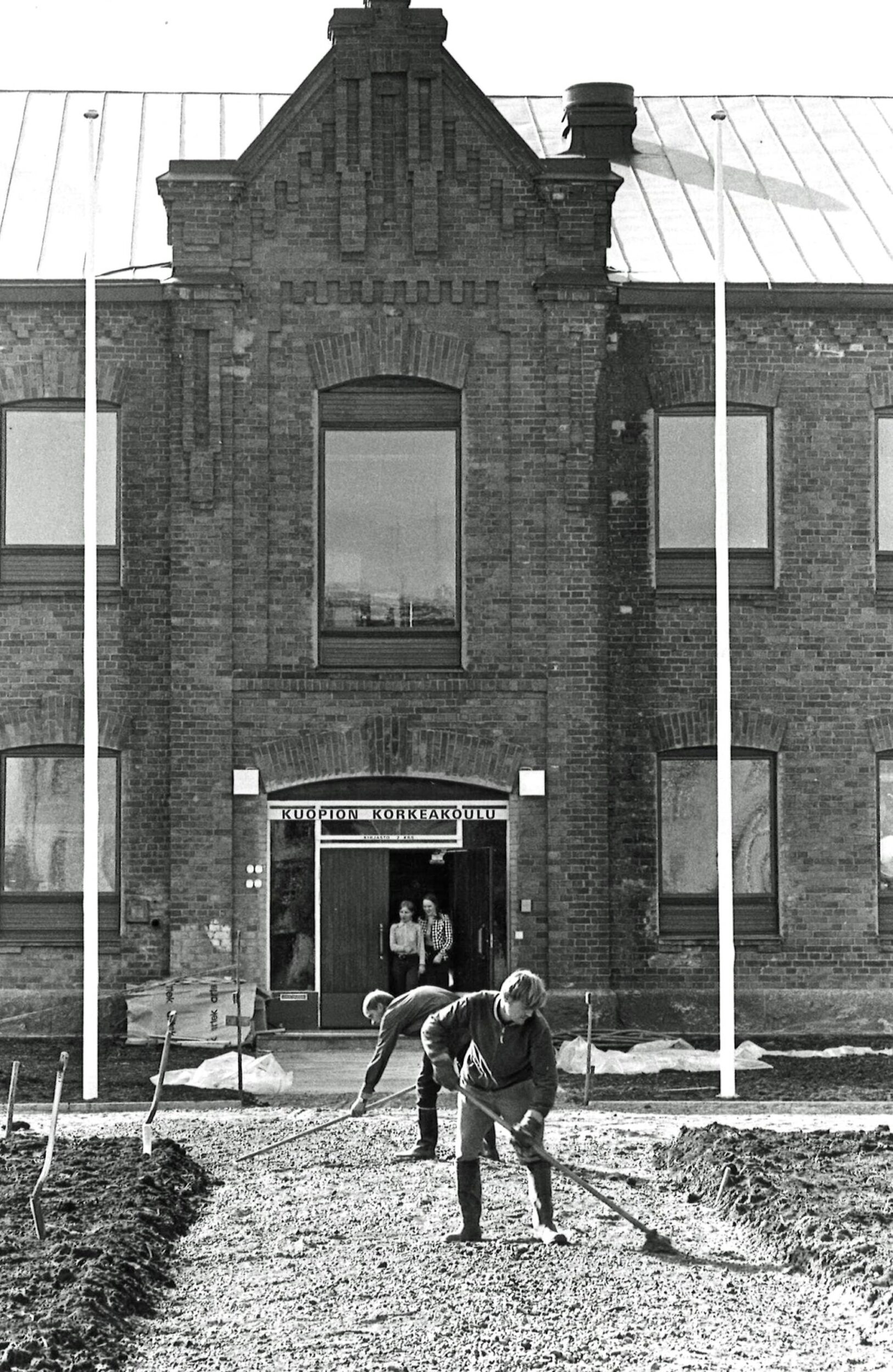Mustavalkoinen kuva tiilirakennukseta, jonka ovesta astuu ulos kaksi henkilöä, ja jonka edustalla kaksi muuta tasoittaa kävelytietä. Lapioita pystyssä etualalla näkyvällä mulloksella. Rakennuksen edessä maassa rakennustarvikkeita. 