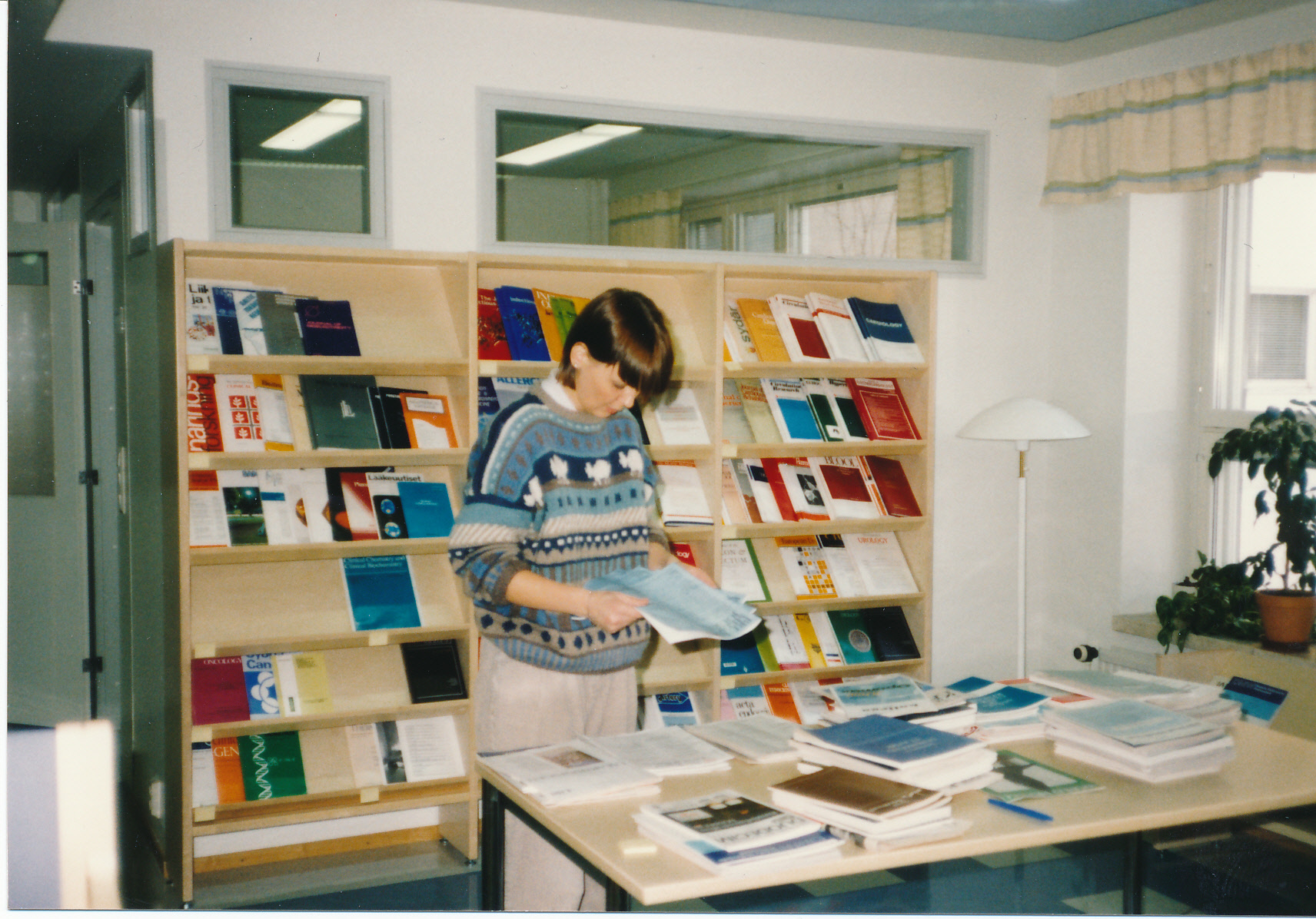 Lehtihylly jossa tieteellisiä lehtiä, pöytä jolla lehtiä, kirjaston työntekijä lajittelee lehtiä.