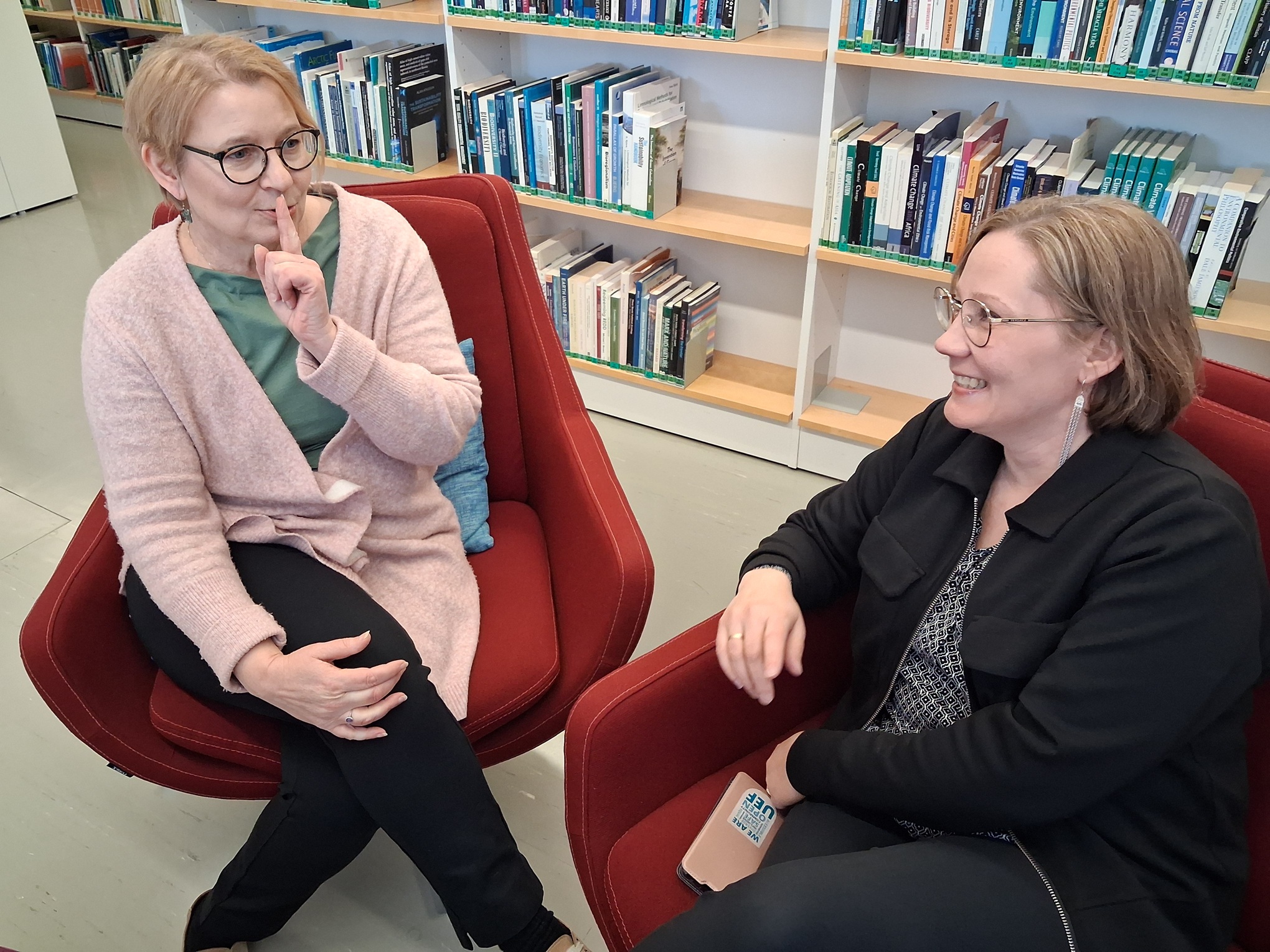 Kaksi naista istuu nojatuolissa. Takana kirjahylly.