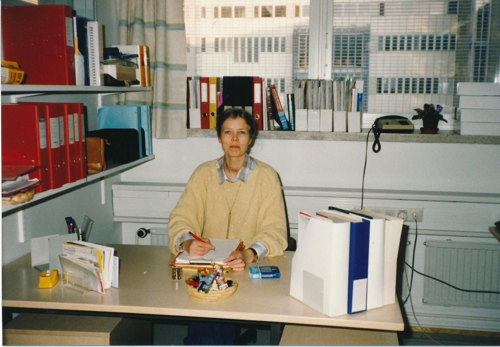 Nainen istuu työpöydän ääressä ja kirjoittaa kynällä paperille. Pöydällä mappeja, koteloita, toimistotarvikkeita, taustalla hylly, jolla mappeja, sekä ikkuna ja ikkunalauta, jolla mappeja, kirjoja, koteloita, lankapuhelin, kasvi. Ikkunasta näkyy toisen rakennuksen seinä.