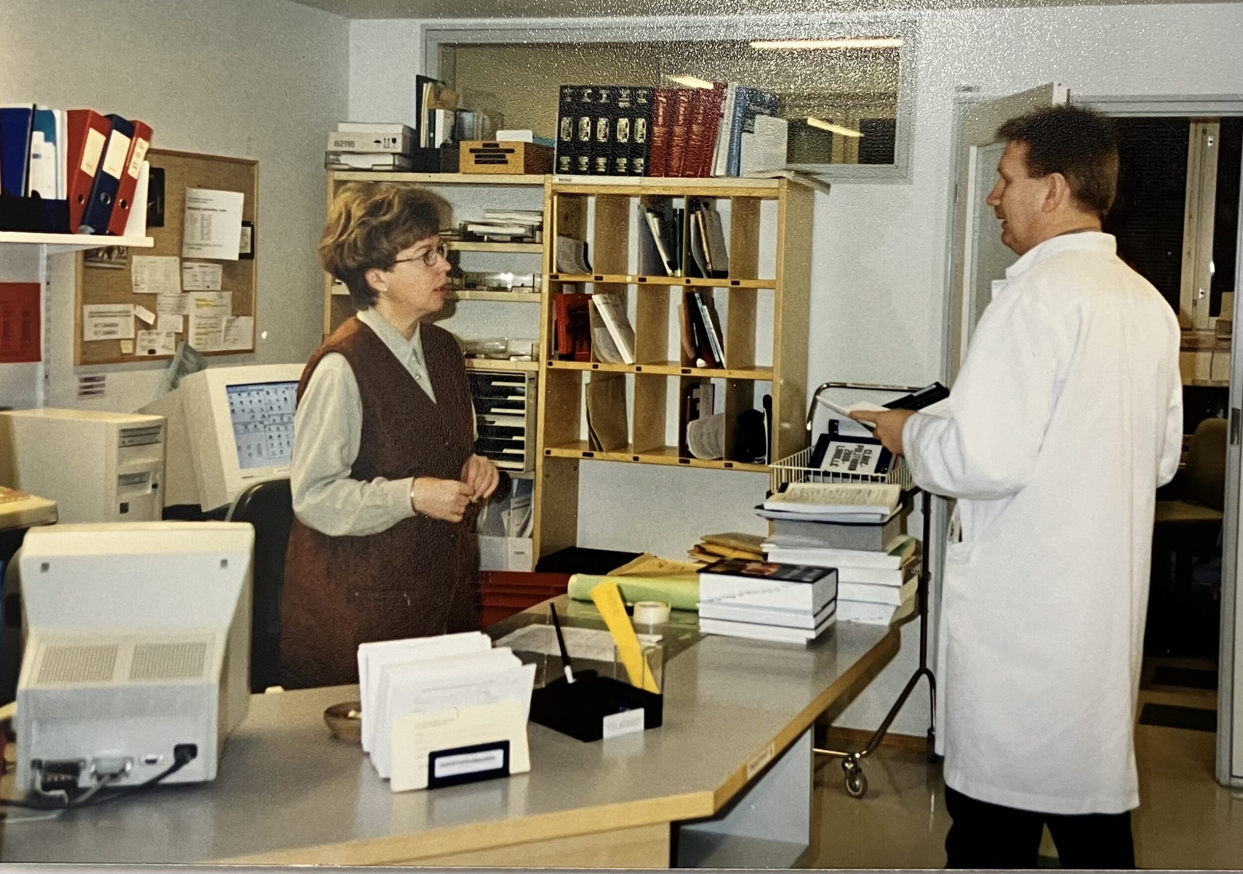 Asiakaspalvelutilanne kirjaston palvelupisteessä. Kaksi ihmistä, joista asiakkaalla valkoinen takki. Kirjoja, hyllyjä, lokeroita, lehitä, tietokoneen kuvaputkinäyttöjä, pöytätietokone, kirjavaunu, ilmoitustaulu, kansioita ja mappeja.