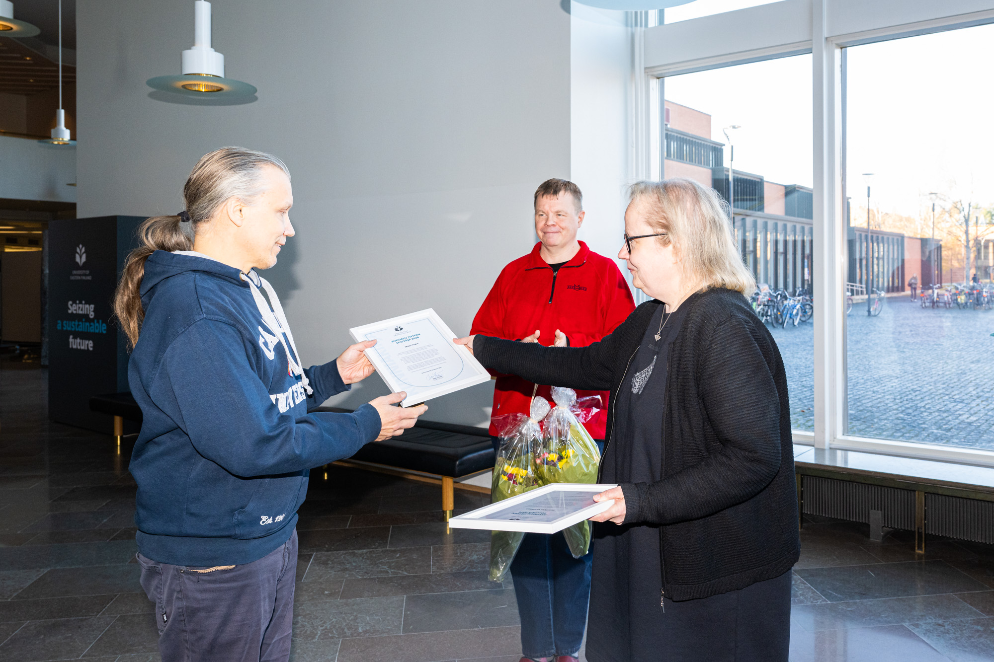 Kolme henkilöä, joista yksi ojentaa toiselle kehystettyä kunniakirjaa ja kolmas seisoo taustalla pidellen kahta kukkapakettia. Three persons, one handing out a framed certificate to the other, and the third standing in the background holding two packages of flowers.