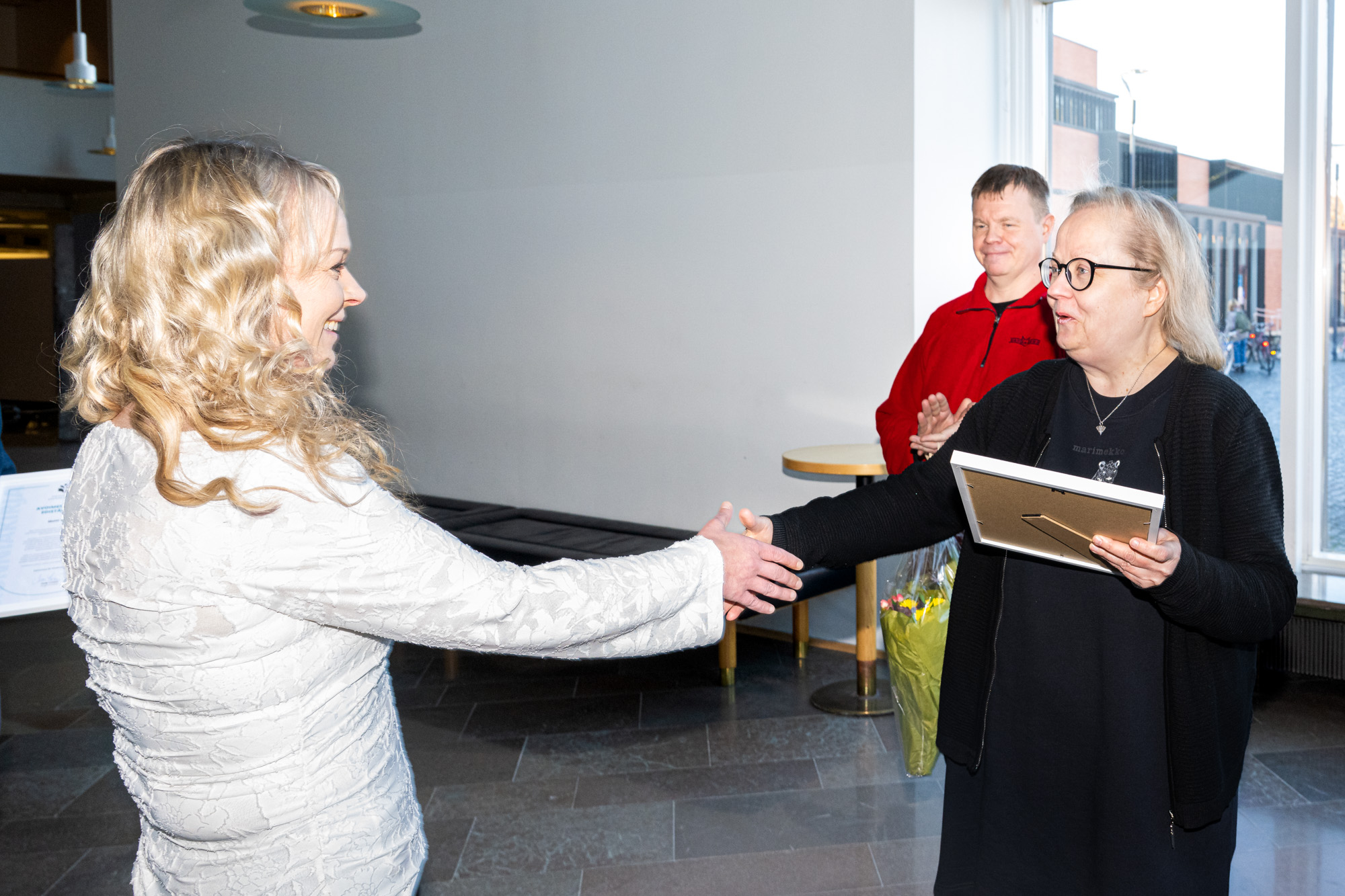 Kaksi henkilöä kättelee. Taustalla kolmas henkilö taputtaa käsiään. Two people shake hands. In the background, a third person claps his hands.