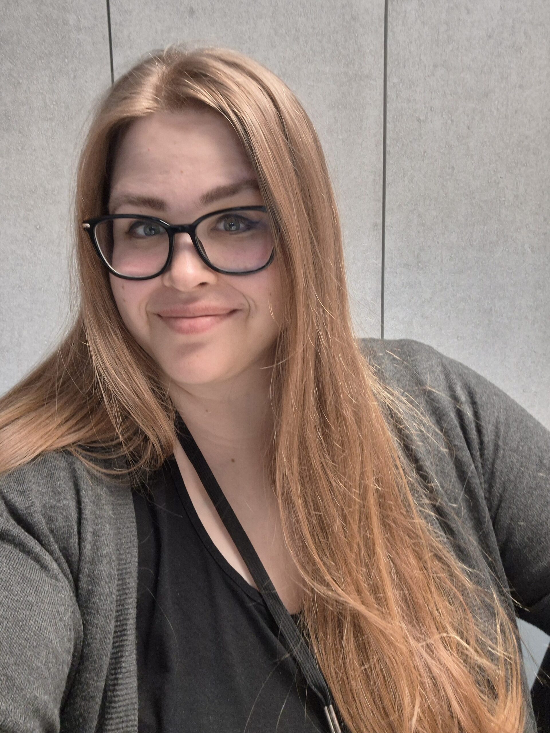 Hymyilevä nainen, jolla on silmälasit ja vaaleanruskeat hiukset. Taustalla on harmaa akustiikkalevy. A smiling woman with eyeglasses and light brown hair. Behind her is a big grey acoustic wall.
